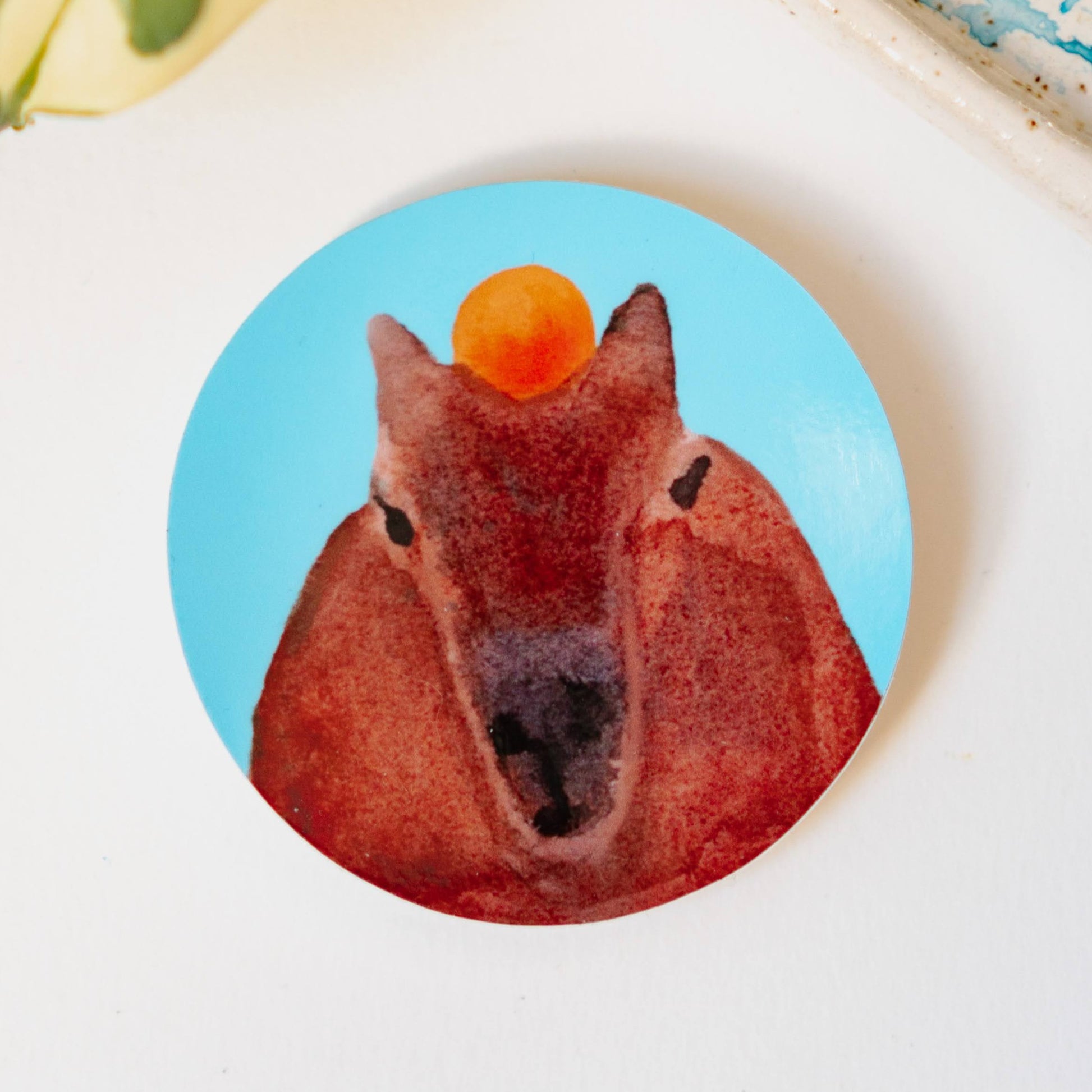A round sticker of a capybara head on a light blue background, facing the viewer with an orange resting on its head.