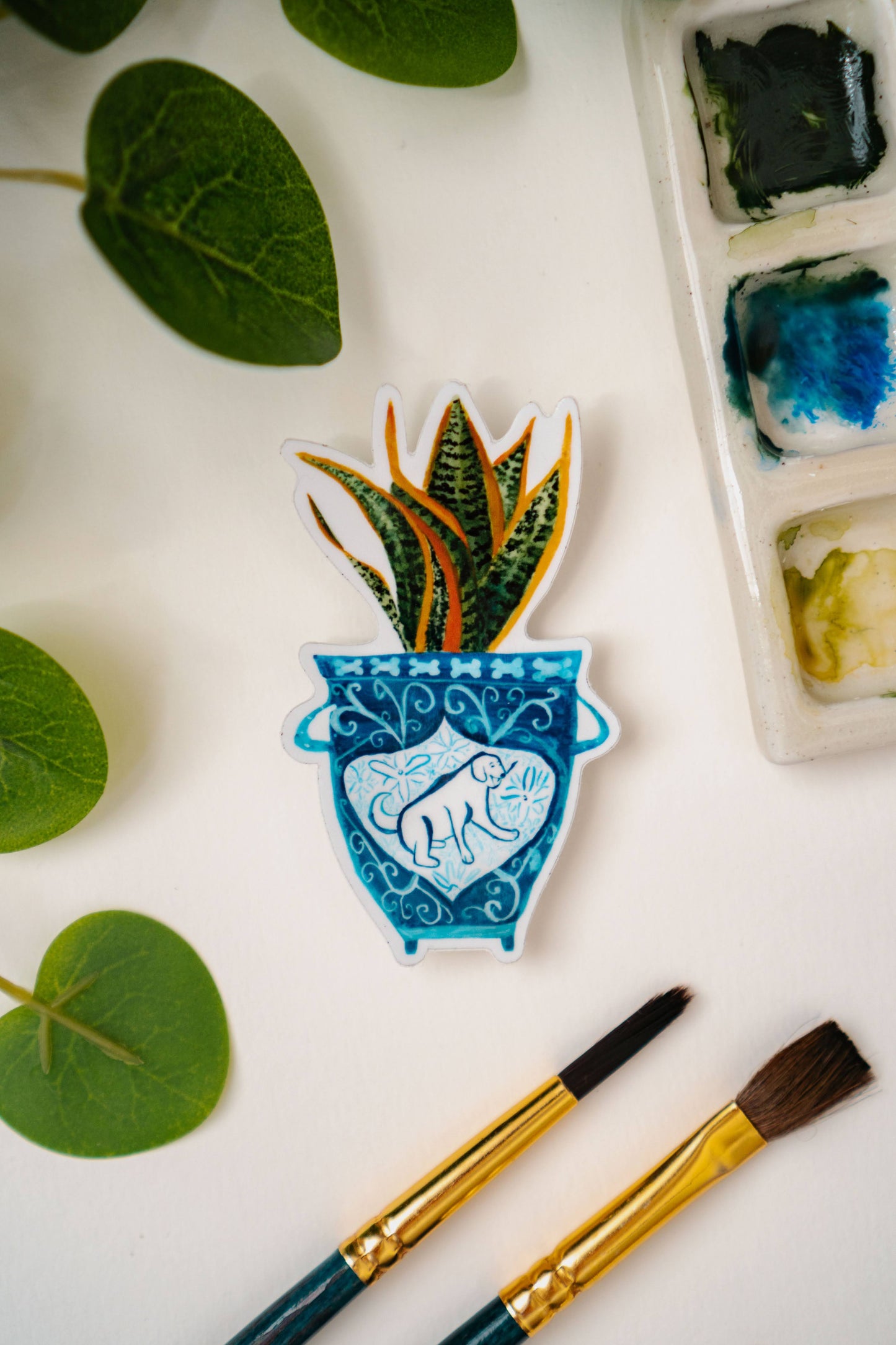 A sticker of a blue and white vase with a dog on it. The vase contains a snake plant.