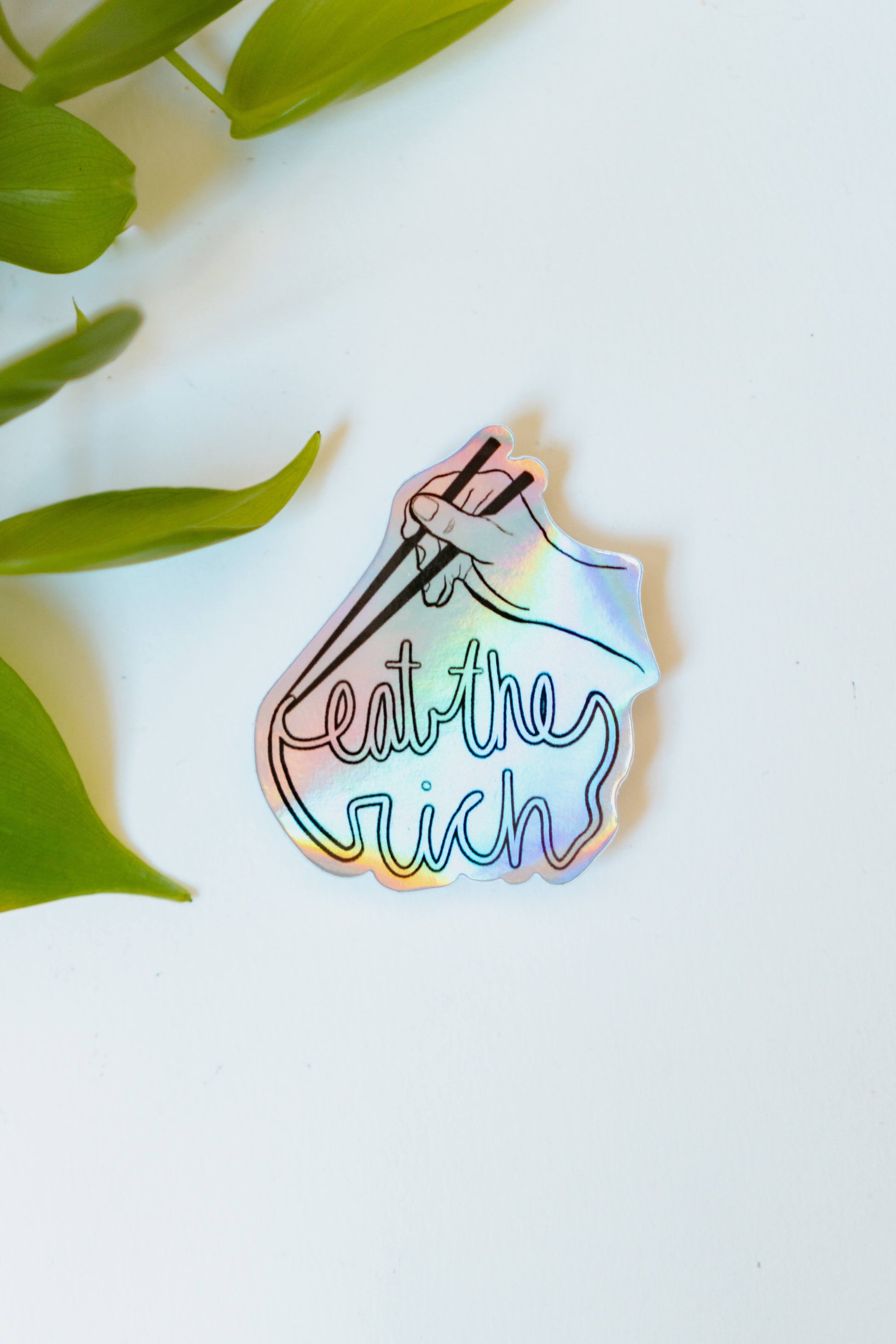 A holographic sticker featuring someone with a pair of chopsticks picking up a noodle which spells, "eat the rich."