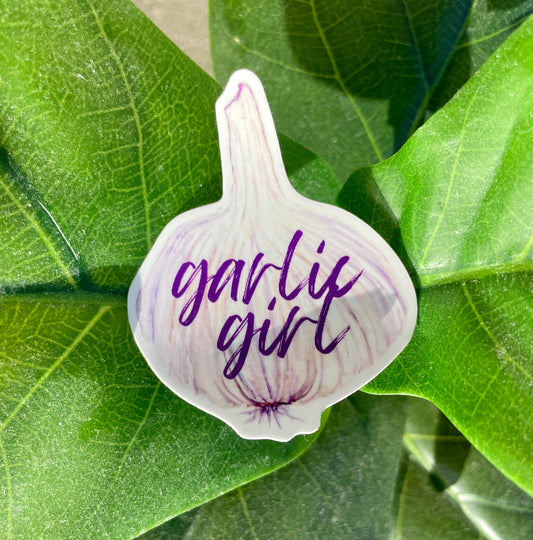 A sticker of a light purple and white garlic bulb with the phrase "garlic girl" written in purple cursive letters.
