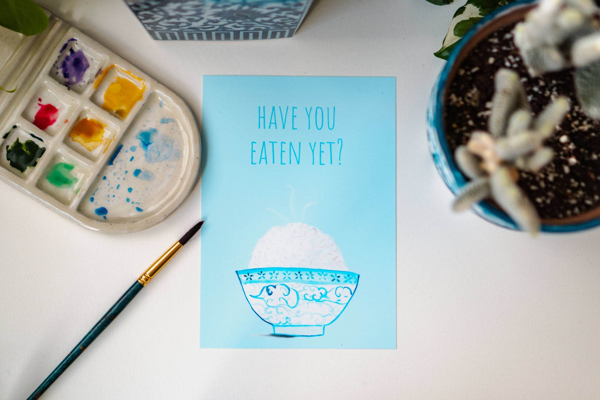 A light blue and white asian art print with the phrase, "Have you eaten yet?" written across the top. A steaming bowl of rice in a white and blue decorated bowl rests at the bottom of the print.