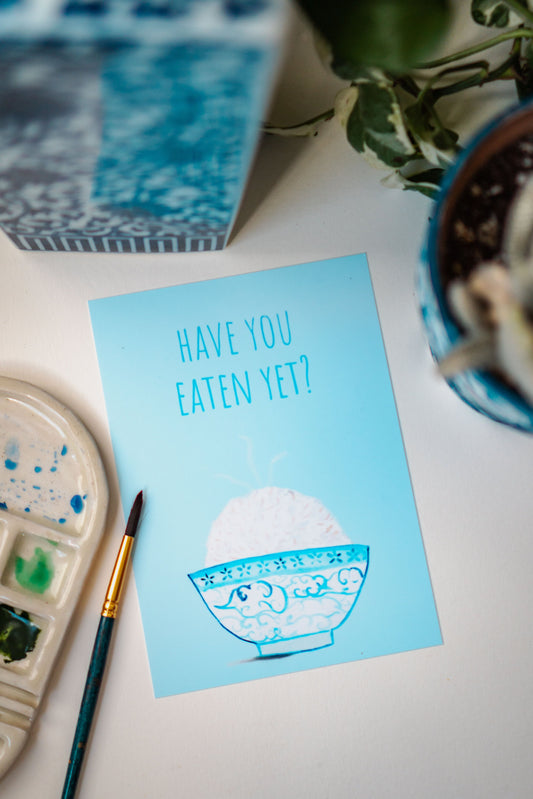 A light blue and white asian art print with the phrase, "Have you eaten yet?" written across the top. A steaming bowl of rice in a white and blue decorated bowl rests at the bottom of the print.
