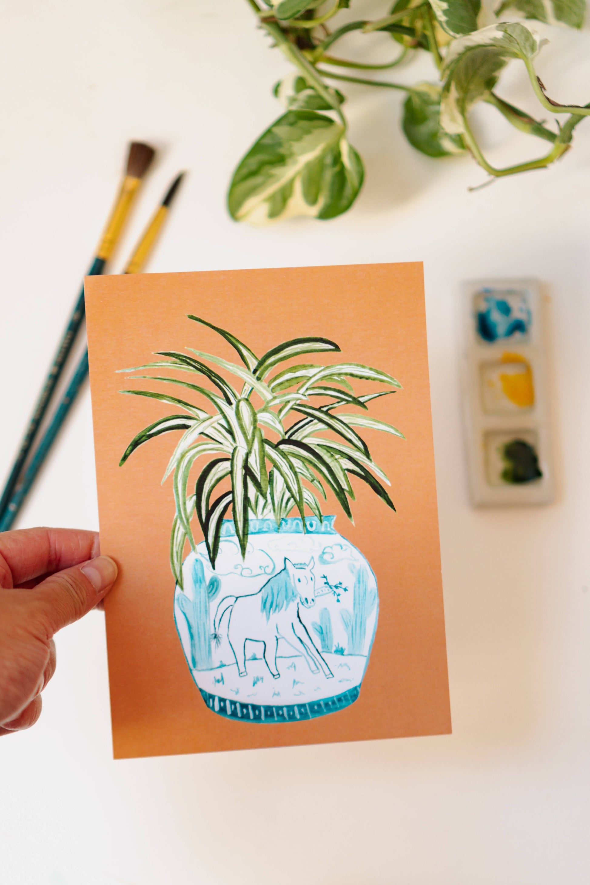 A print of a spider plant in a blue and white porcelain vase, decorated with a horse.