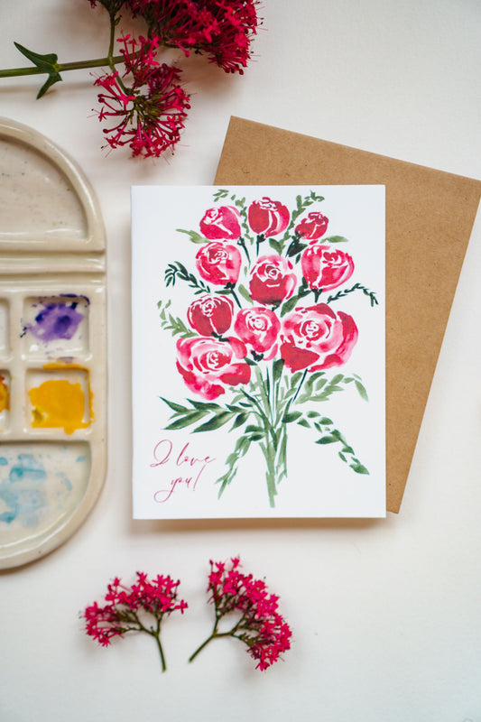 A white greeting card with red watercolor roses and "I love you!" written in thin, cursive script in the bottom left corner.