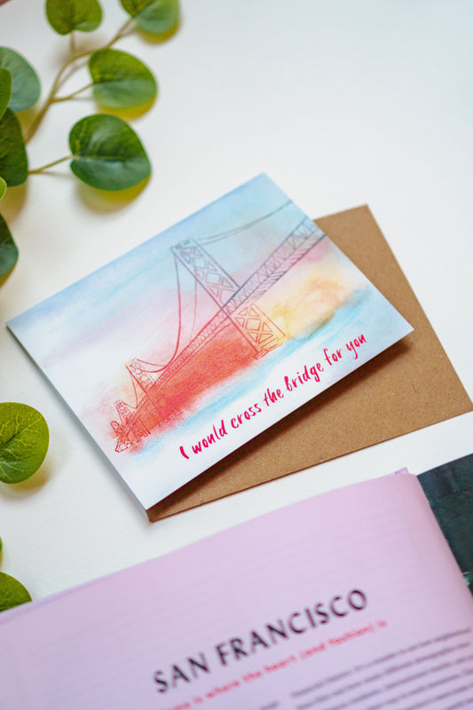 A card with a watercolor rendition of the San Francisco Bay Bridge on it, painted in oranges, yellows, and blues. The phrase "I would cross the bridge for you" is written on the bottom of the card.