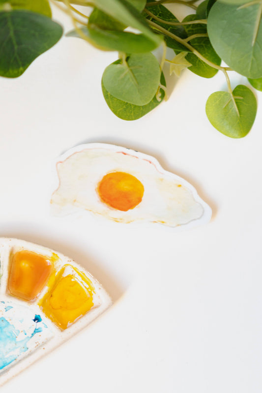 A narrow, white sticker of a fried, sunny side up egg with a rich yellow yolk and crispy edges.