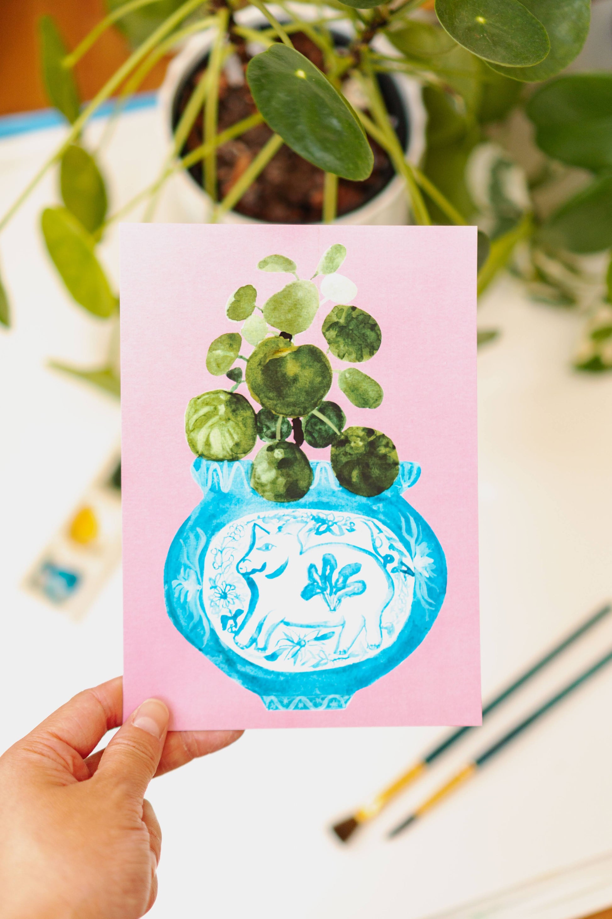 A print of a pilea plant in a blue and white porcelain vase, decorated with a pig.