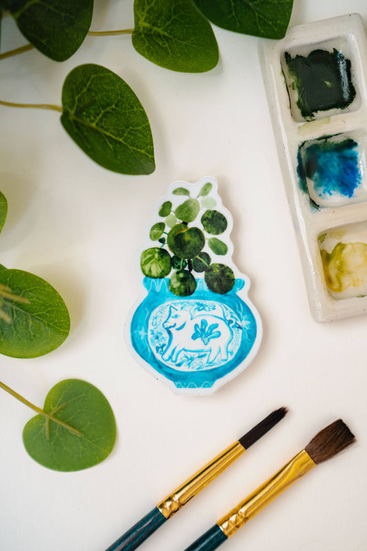A sticker of a pilea plant in a blue and white porcelain vase, decorated with a pig.