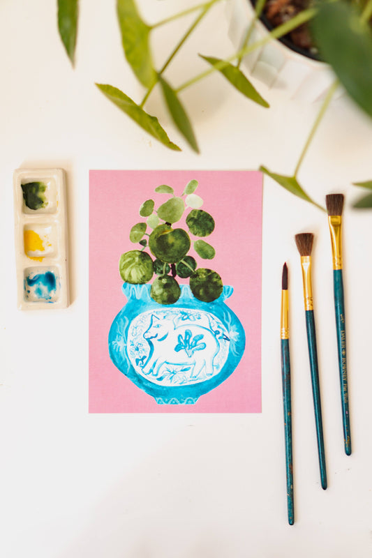 A print of a pilea plant in a blue and white porcelain vase, decorated with a pig.