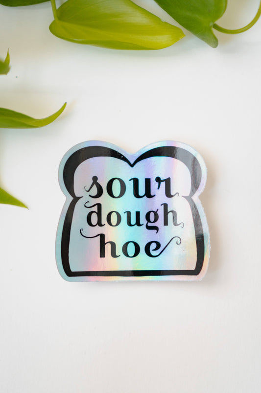 A holographic sticker featuring line art of a slice of bread and the words "sourdough hoe" written in the center.