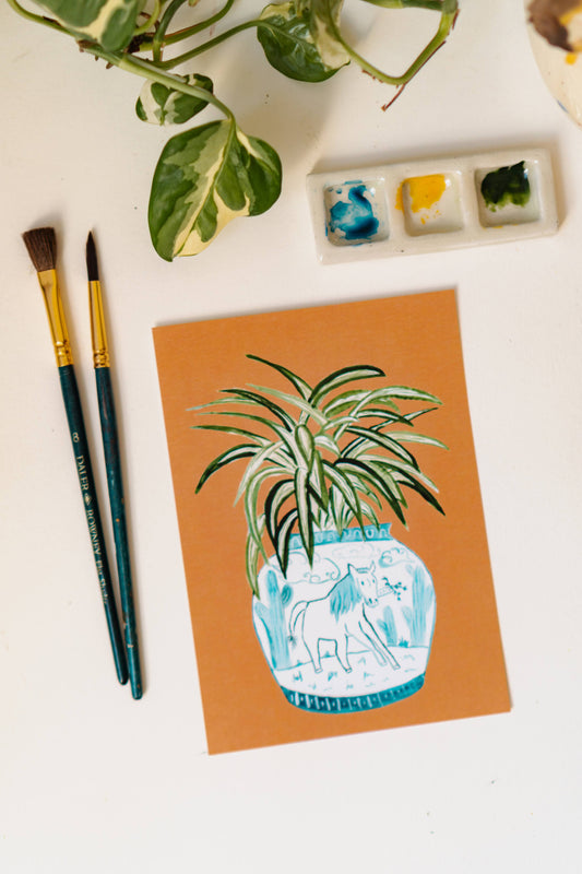 A print of a spider plant in a blue and white porcelain vase, decorated with a horse.