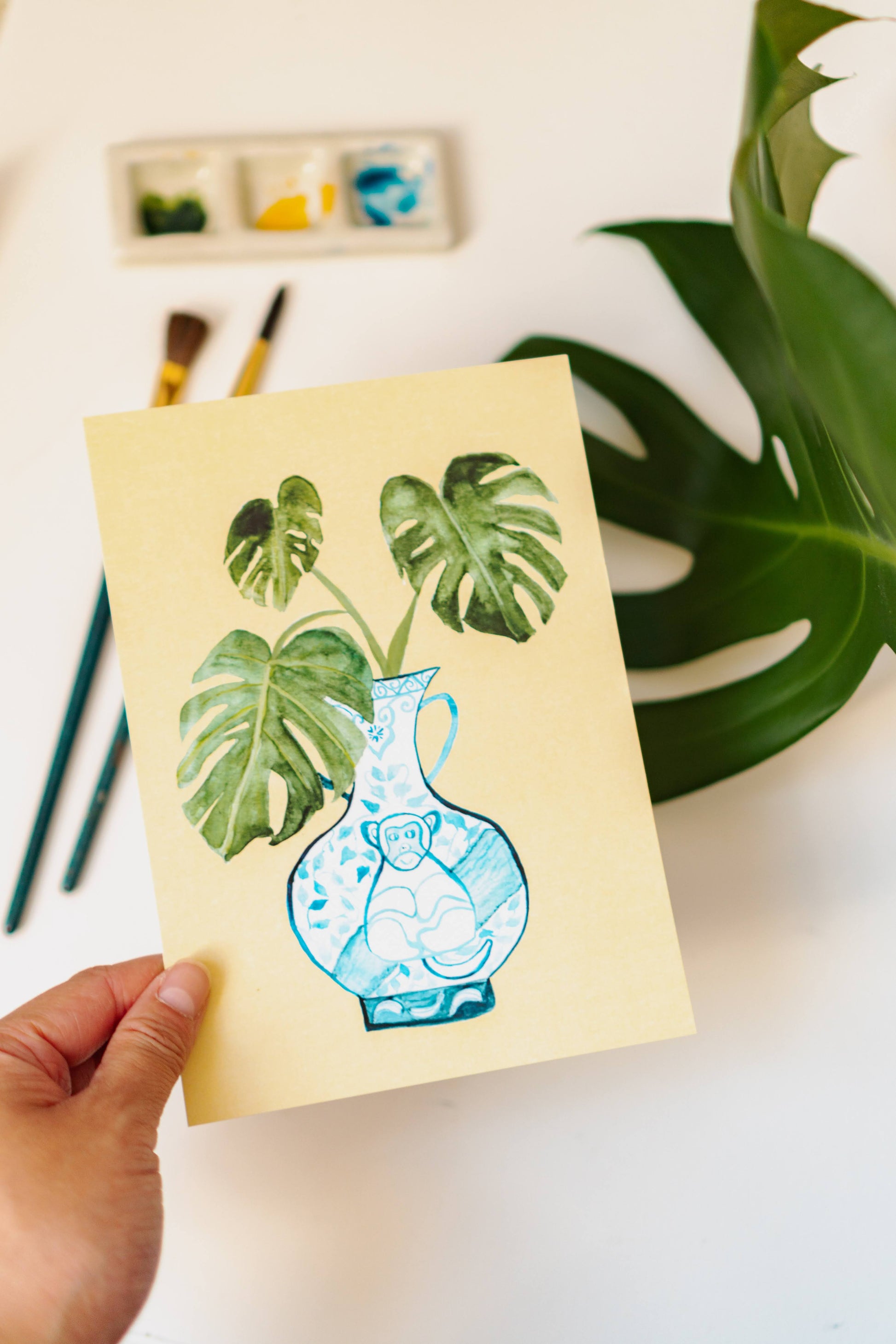 A print of a monstera plant in a blue and white porcelain vase, decorated with a monkey.