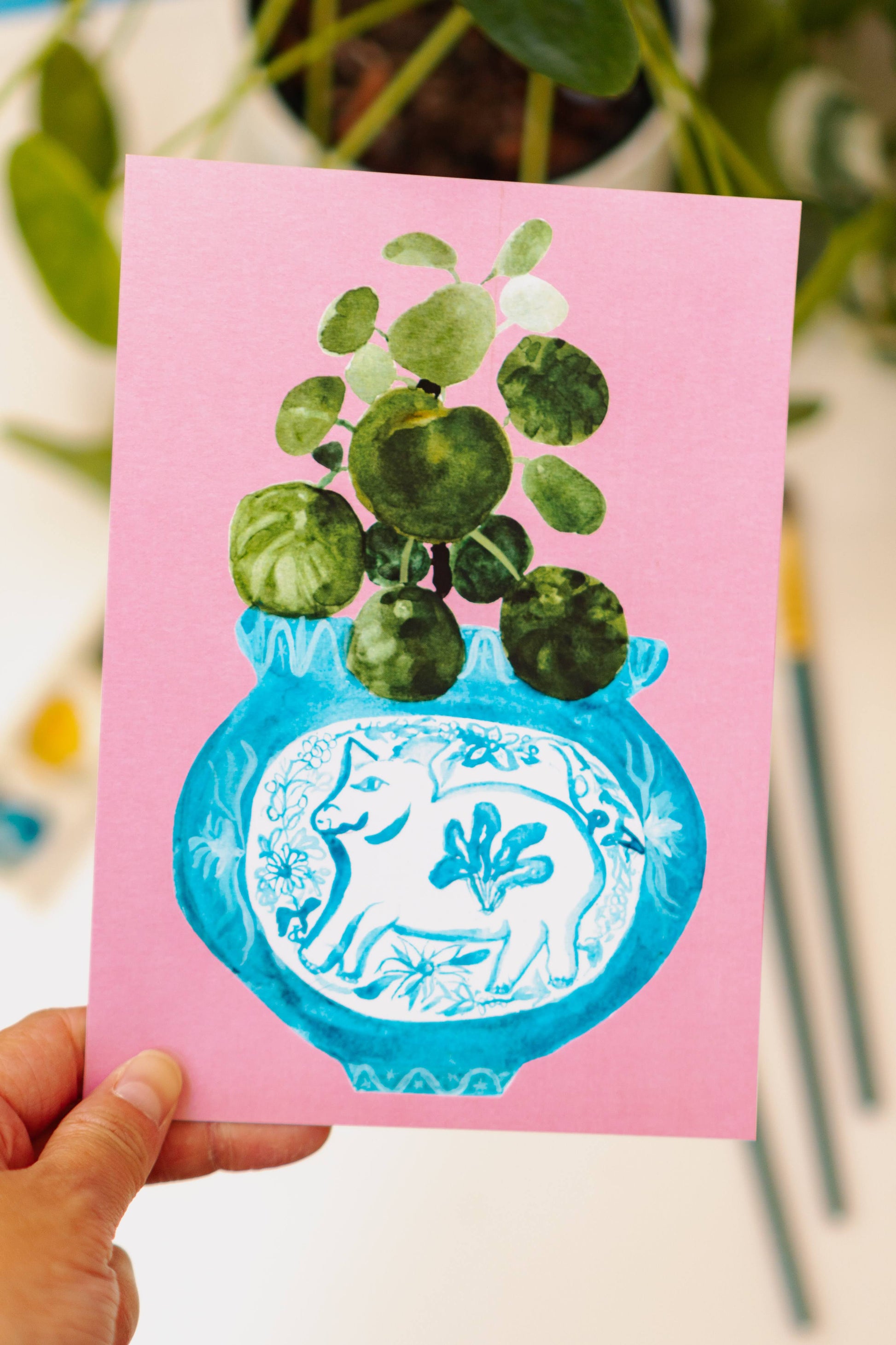 A print of a pilea plant in a blue and white porcelain vase, decorated with a pig.