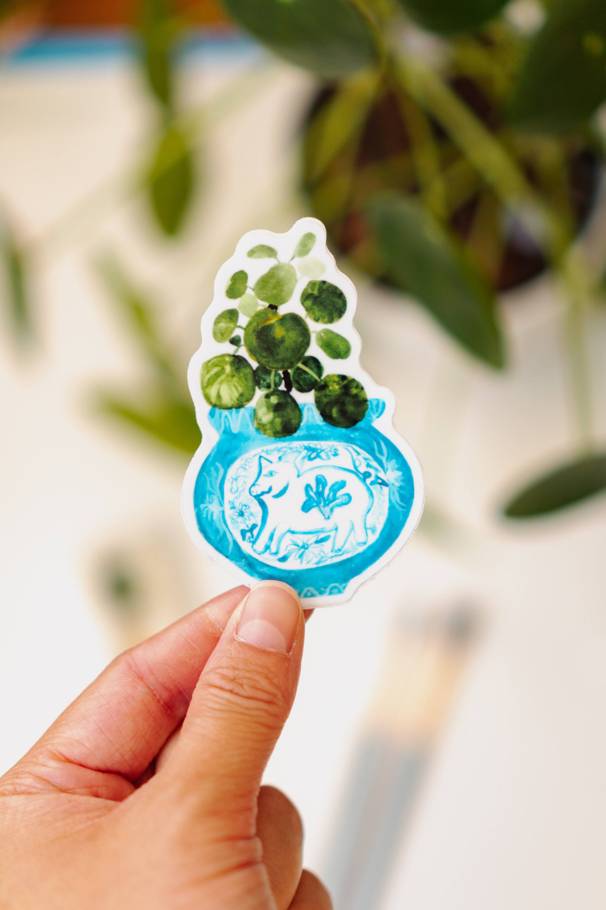 A sticker of a pilea plant in a blue and white porcelain vase, decorated with a pig.