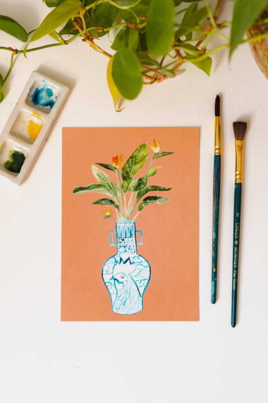 A print of a bird of paradise plant in a blue and white porcelain vase, decorated with a rooster.