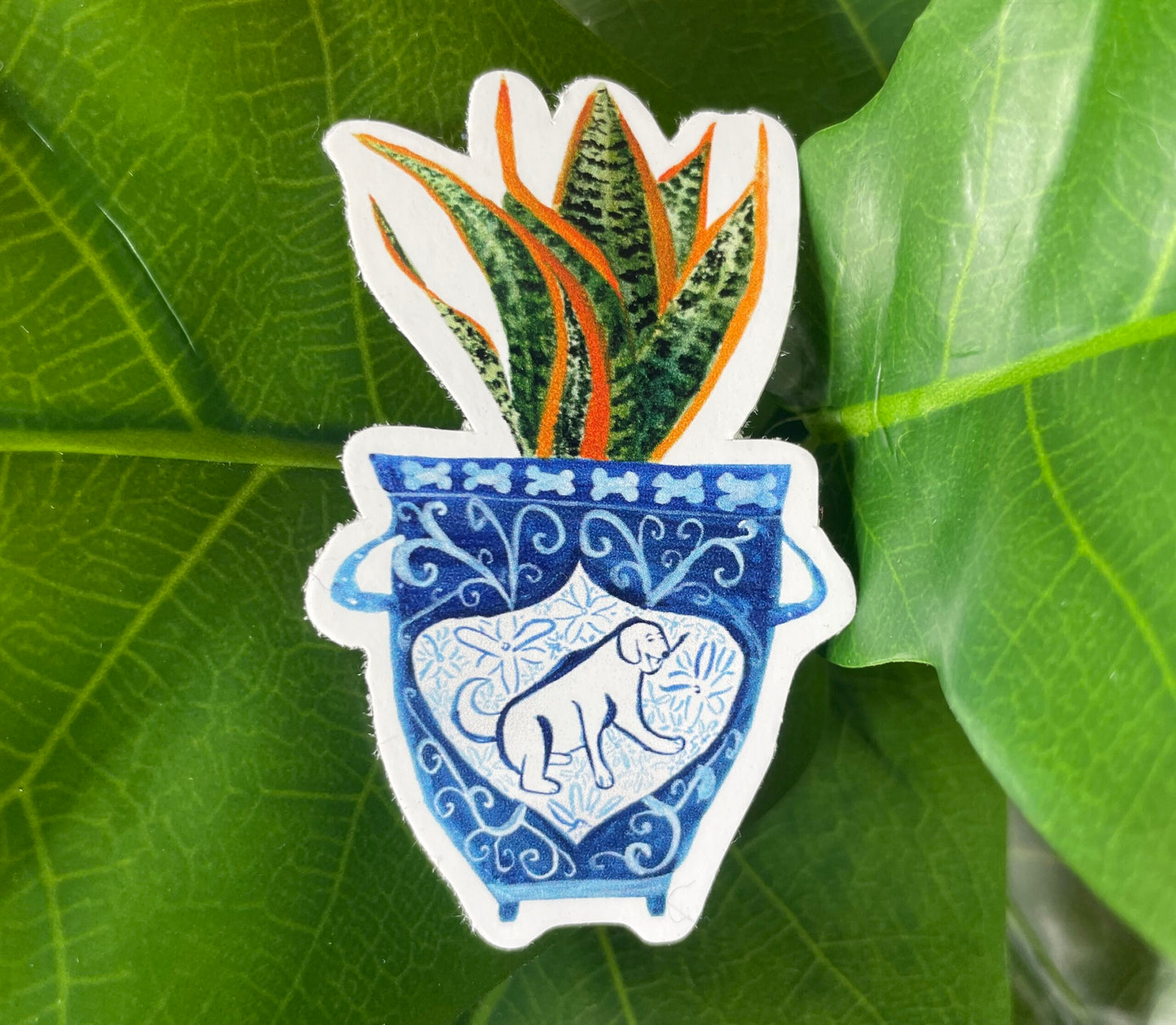 A sticker of a blue and white vase with a dog on it. The vase contains a snake plant.