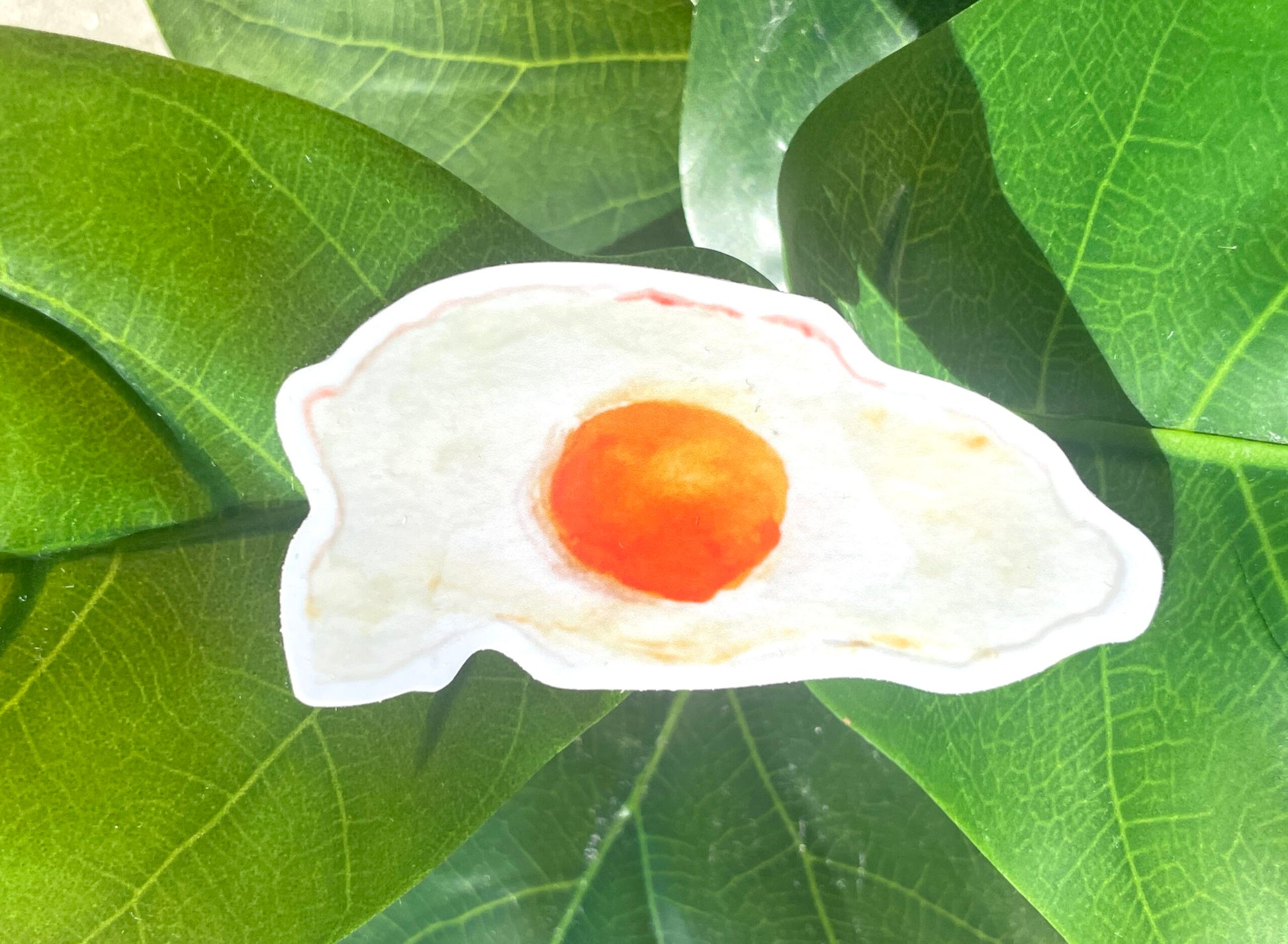 A narrow, white sticker of a fried, sunny side up egg with a rich yellow yolk and crispy edges.