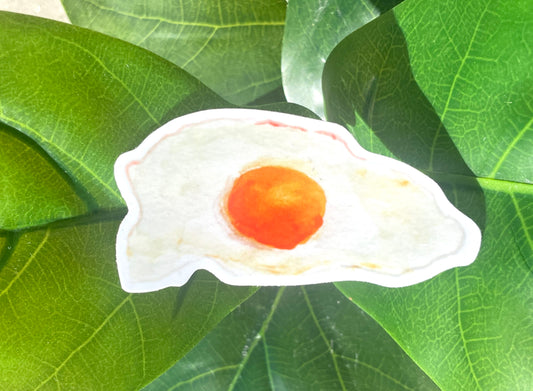 A narrow, white sticker of a fried, sunny side up egg with a rich yellow yolk and crispy edges.