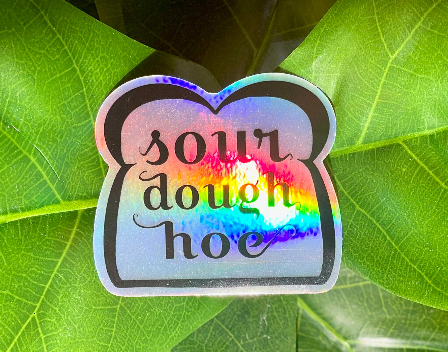 A holographic sticker featuring line art of a slice of bread and the words "sourdough hoe" written in the center.