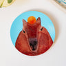 A round sticker of a capybara head on a light blue background, facing the viewer with an orange resting on its head.