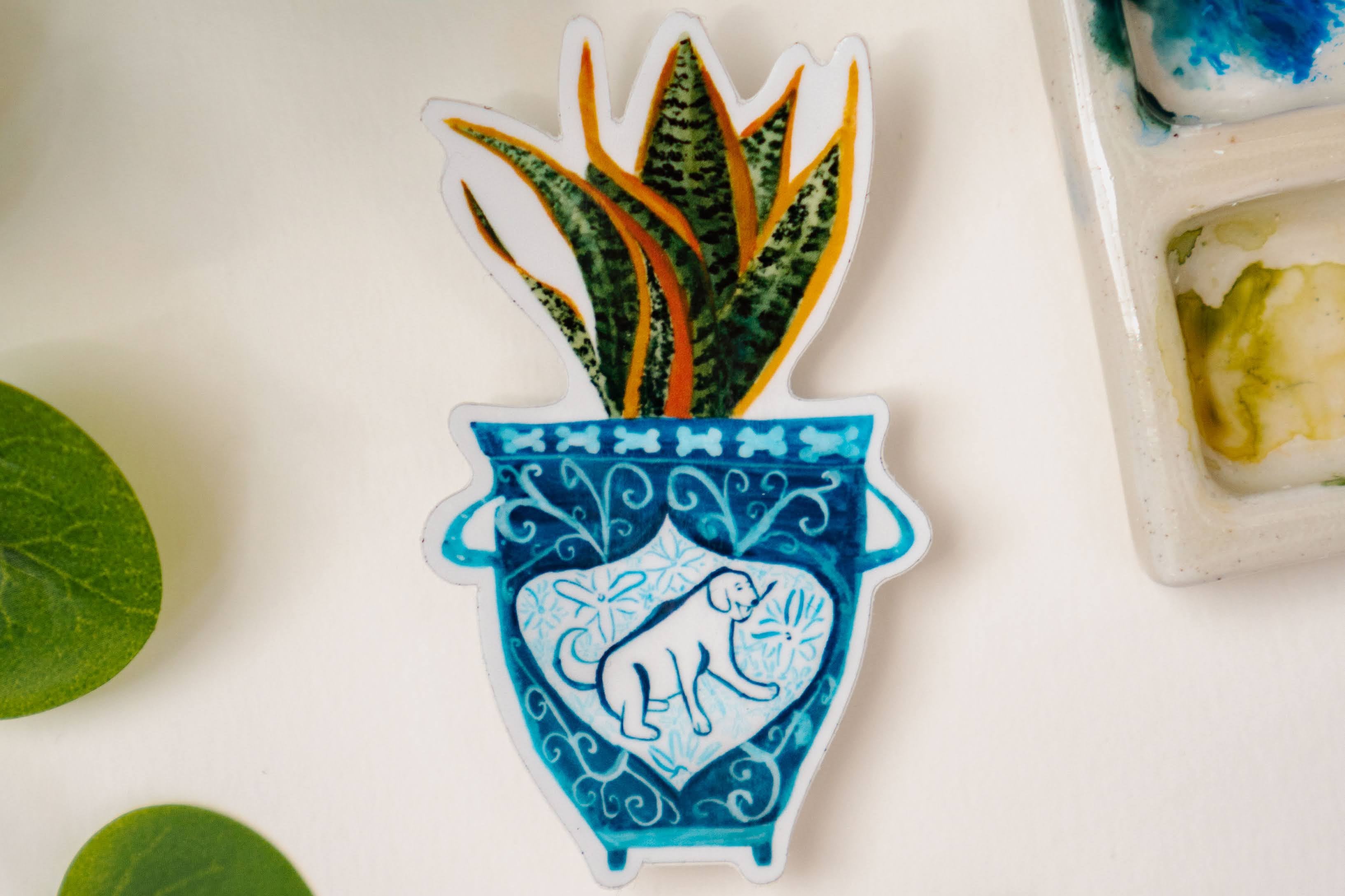 A sticker of a blue and white vase with a dog on it. The vase contains a snake plant.