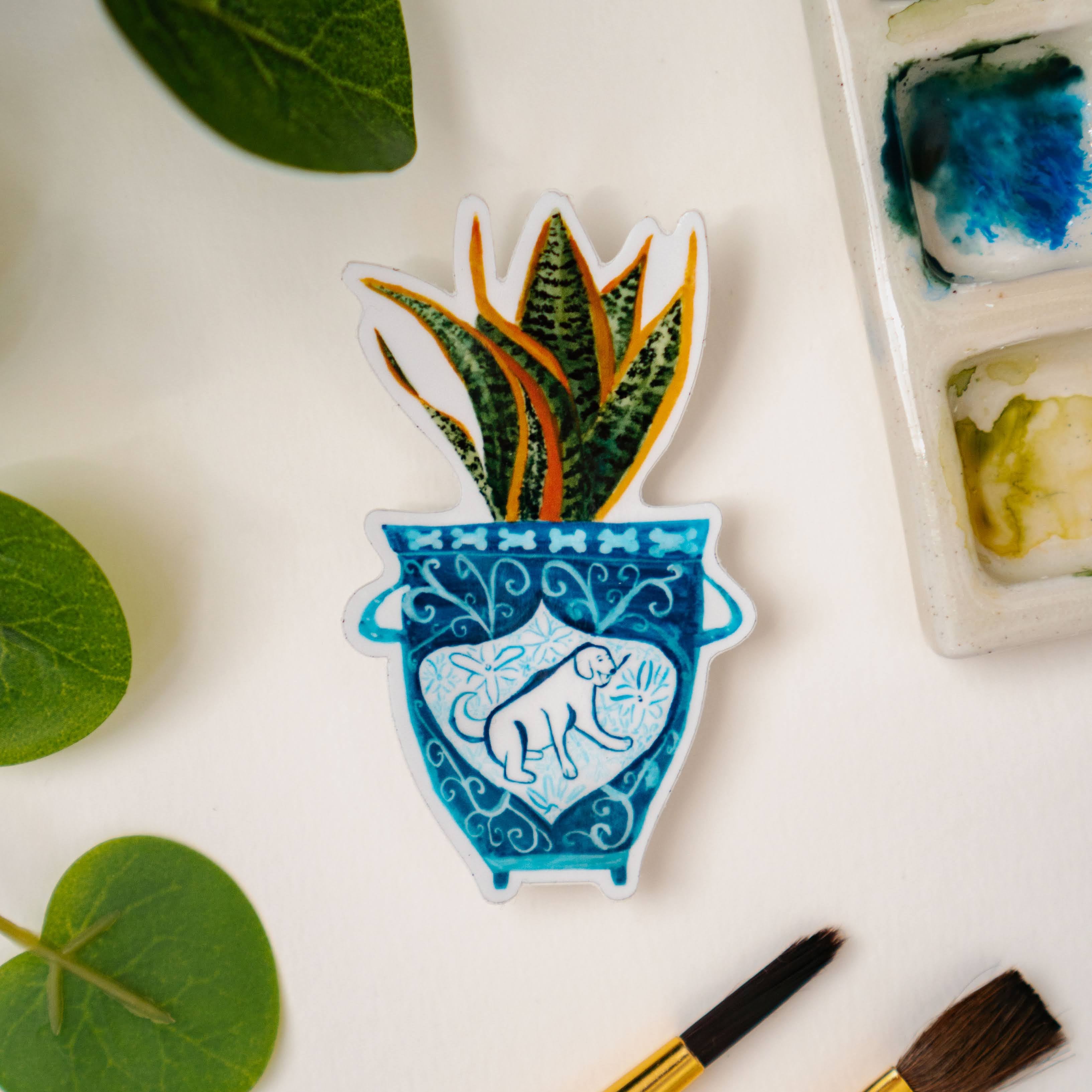 A sticker of a blue and white vase with a dog on it. The vase contains a snake plant.