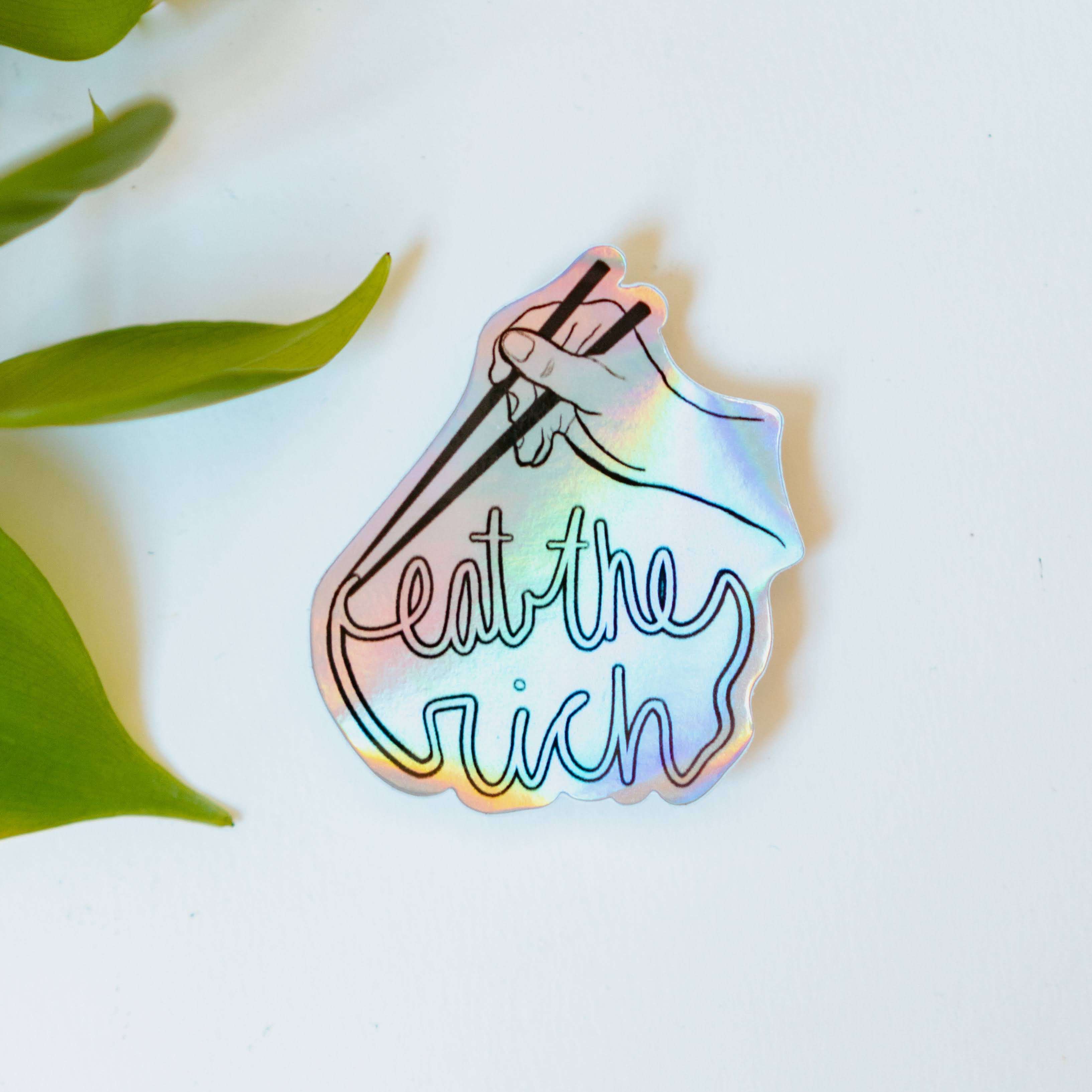 A holographic sticker featuring someone with a pair of chopsticks picking up a noodle which spells, "eat the rich."