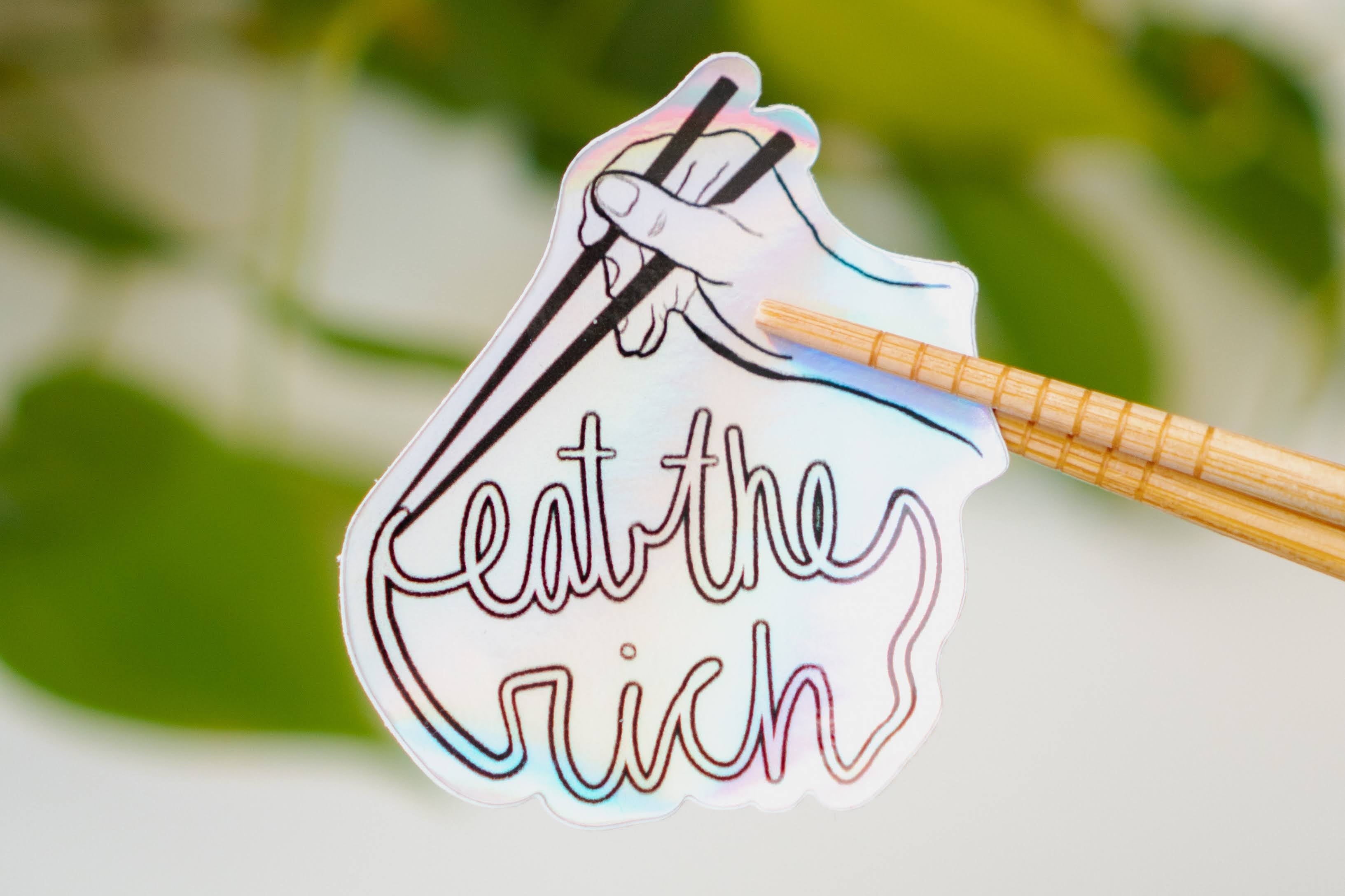 A holographic sticker featuring someone with a pair of chopsticks picking up a noodle which spells, "eat the rich."