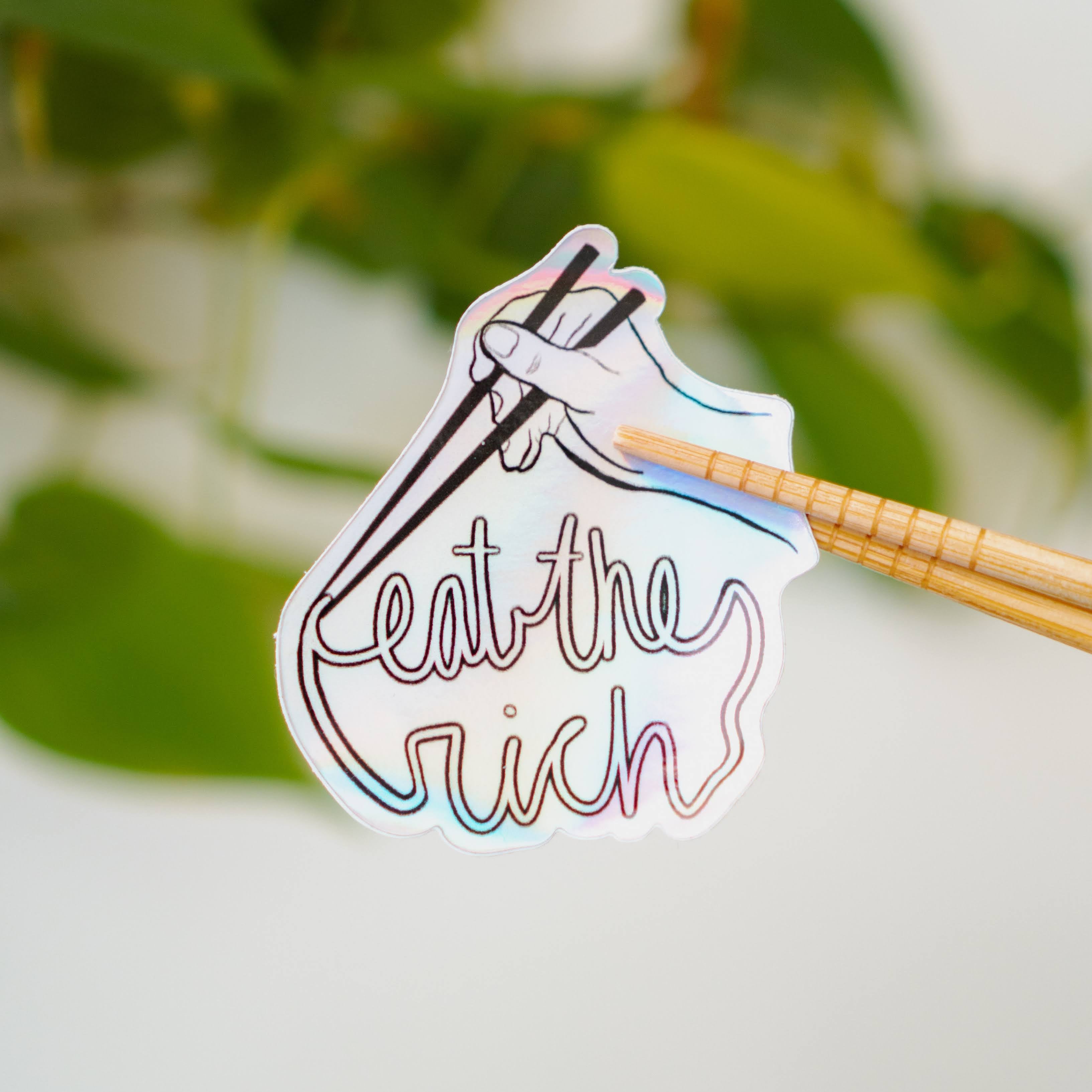 A holographic sticker featuring someone with a pair of chopsticks picking up a noodle which spells, "eat the rich."