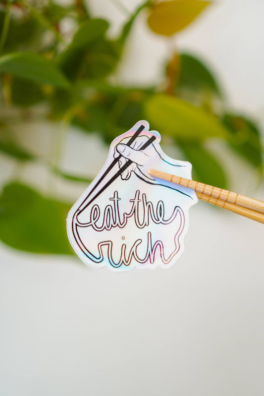 A holographic sticker featuring someone with a pair of chopsticks picking up a noodle which spells, "eat the rich."