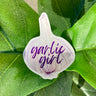 A sticker of a light purple and white garlic bulb with the phrase "garlic girl" written in purple cursive letters.