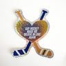 A sparkly, holographic die-cut sticker featuring two crossed hockey sticks. In the center where the sticks cross is a heart shape containing the text "GAY HOCKEY MADE ME BELIEVE IN LOVE AGAIN" in bold white letters