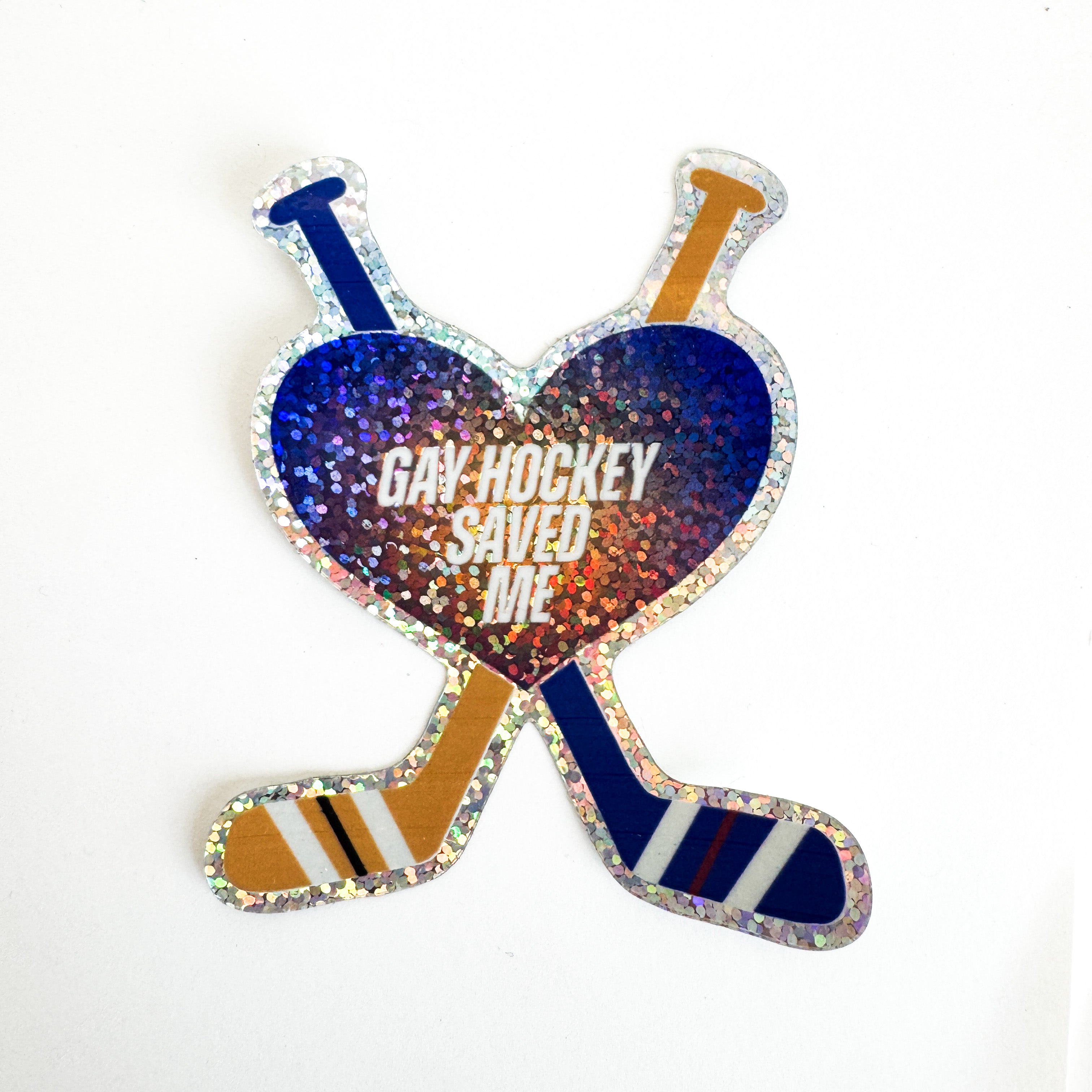 A die-cut sticker featuring a heart layered over two crossed hockey sticks. Bold white text inside the heart reads "GAY HOCKEY SAVED ME." The entire sticker has a shiny, holographic glitter finish.