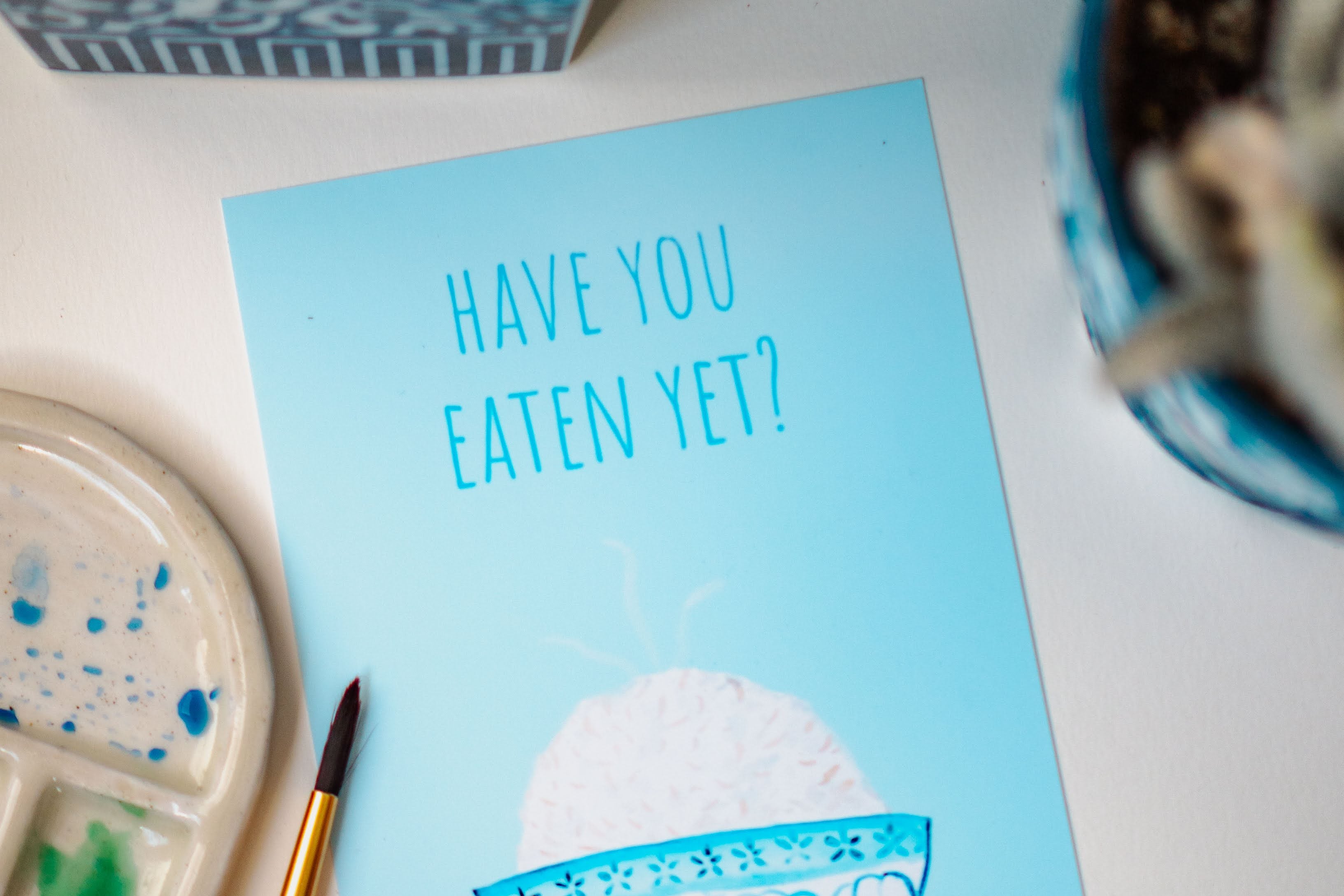 A light blue and white asian art print with the phrase, "Have you eaten yet?" written across the top. A steaming bowl of rice in a white and blue decorated bowl rests at the bottom of the print.