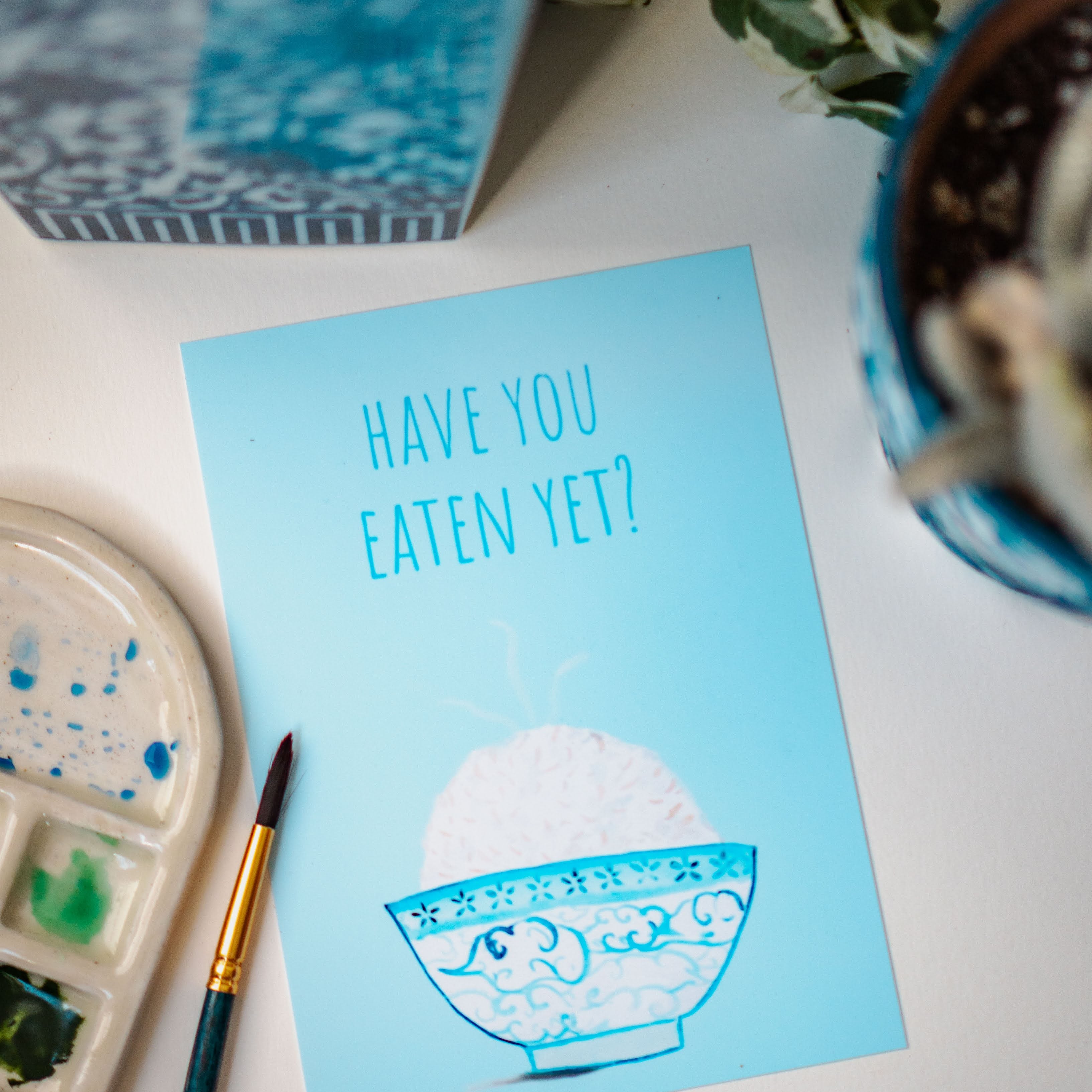 A light blue and white asian art print with the phrase, "Have you eaten yet?" written across the top. A steaming bowl of rice in a white and blue decorated bowl rests at the bottom of the print.