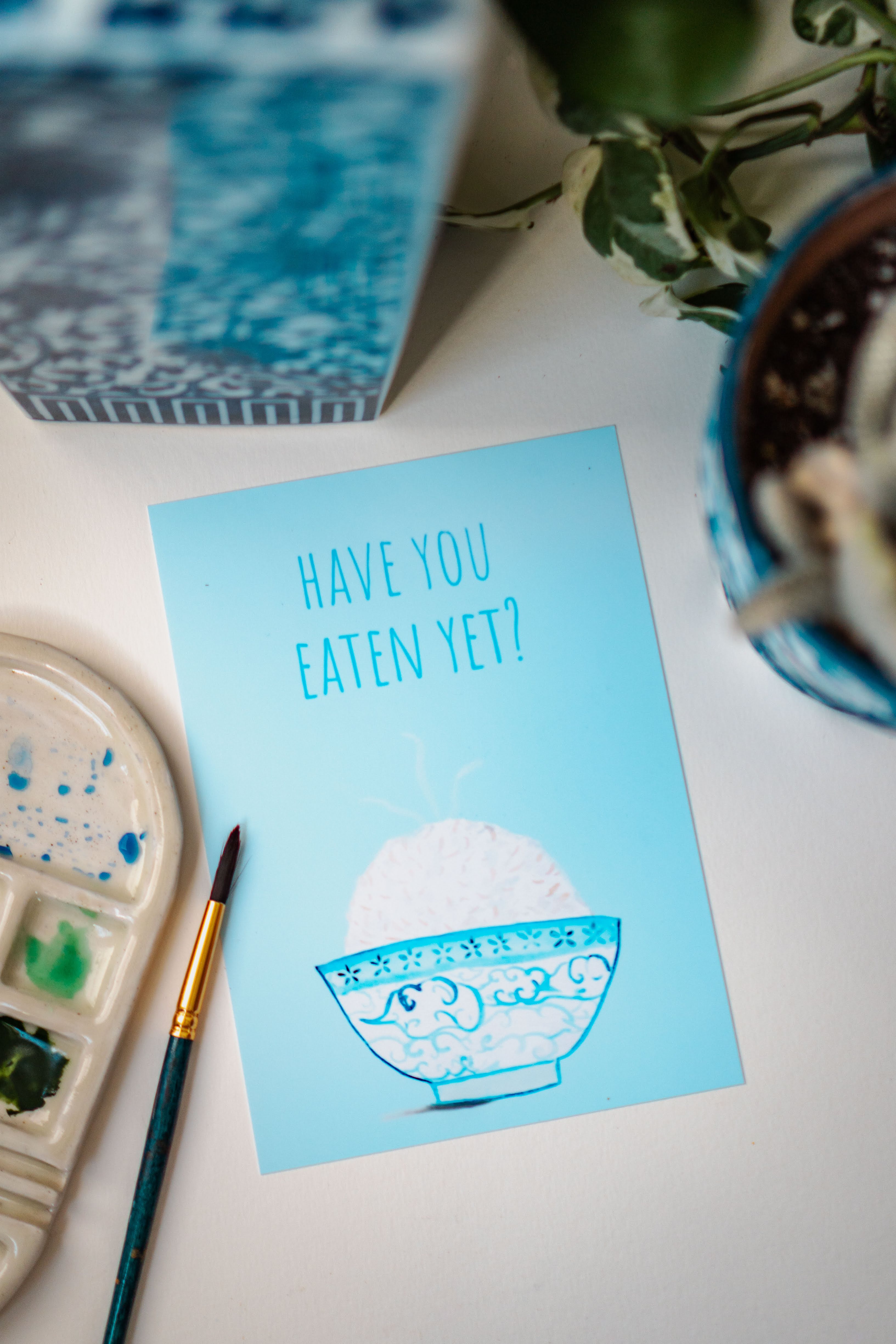 A light blue and white asian art print with the phrase, "Have you eaten yet?" written across the top. A steaming bowl of rice in a white and blue decorated bowl rests at the bottom of the print.