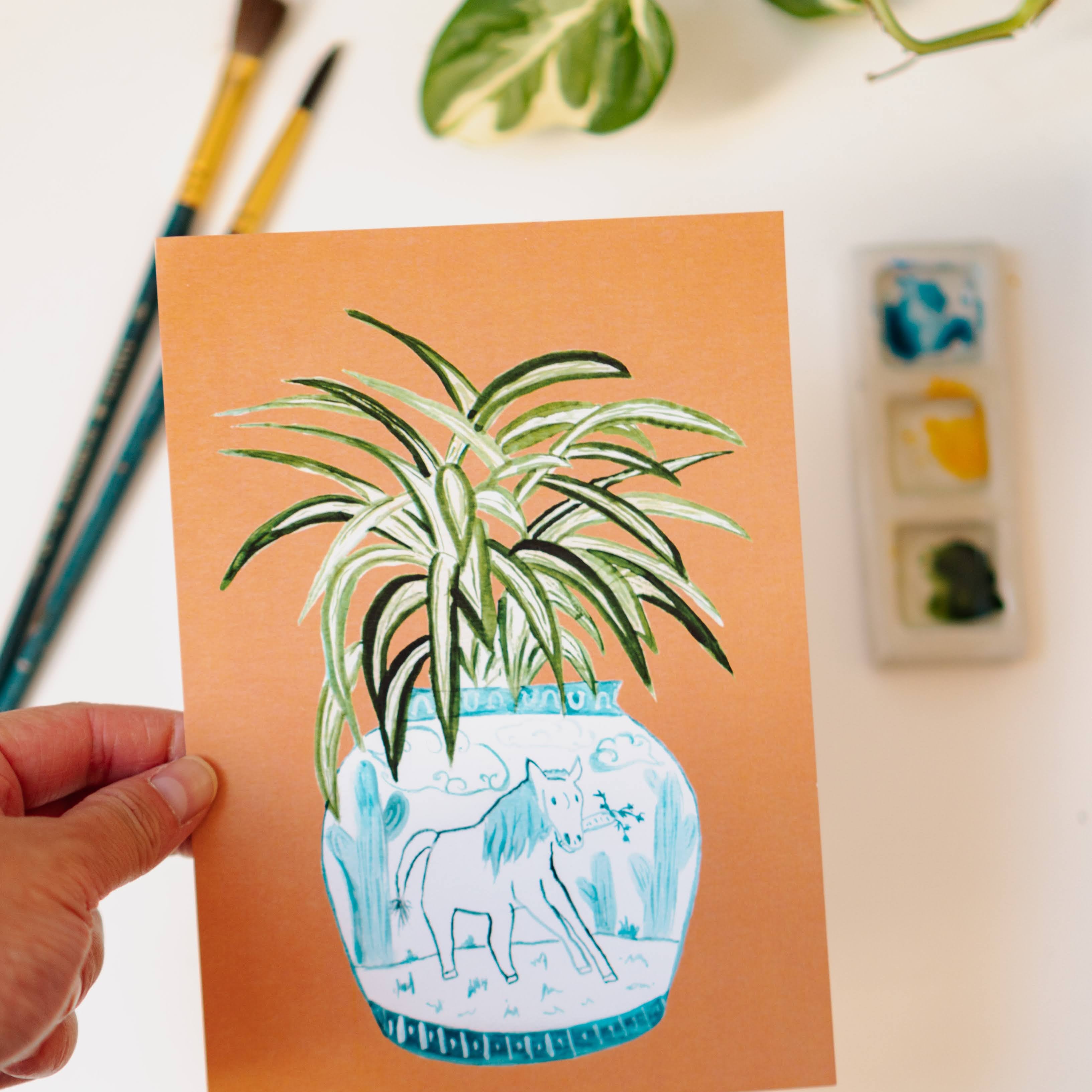 A print of a spider plant in a blue and white porcelain vase, decorated with a horse.