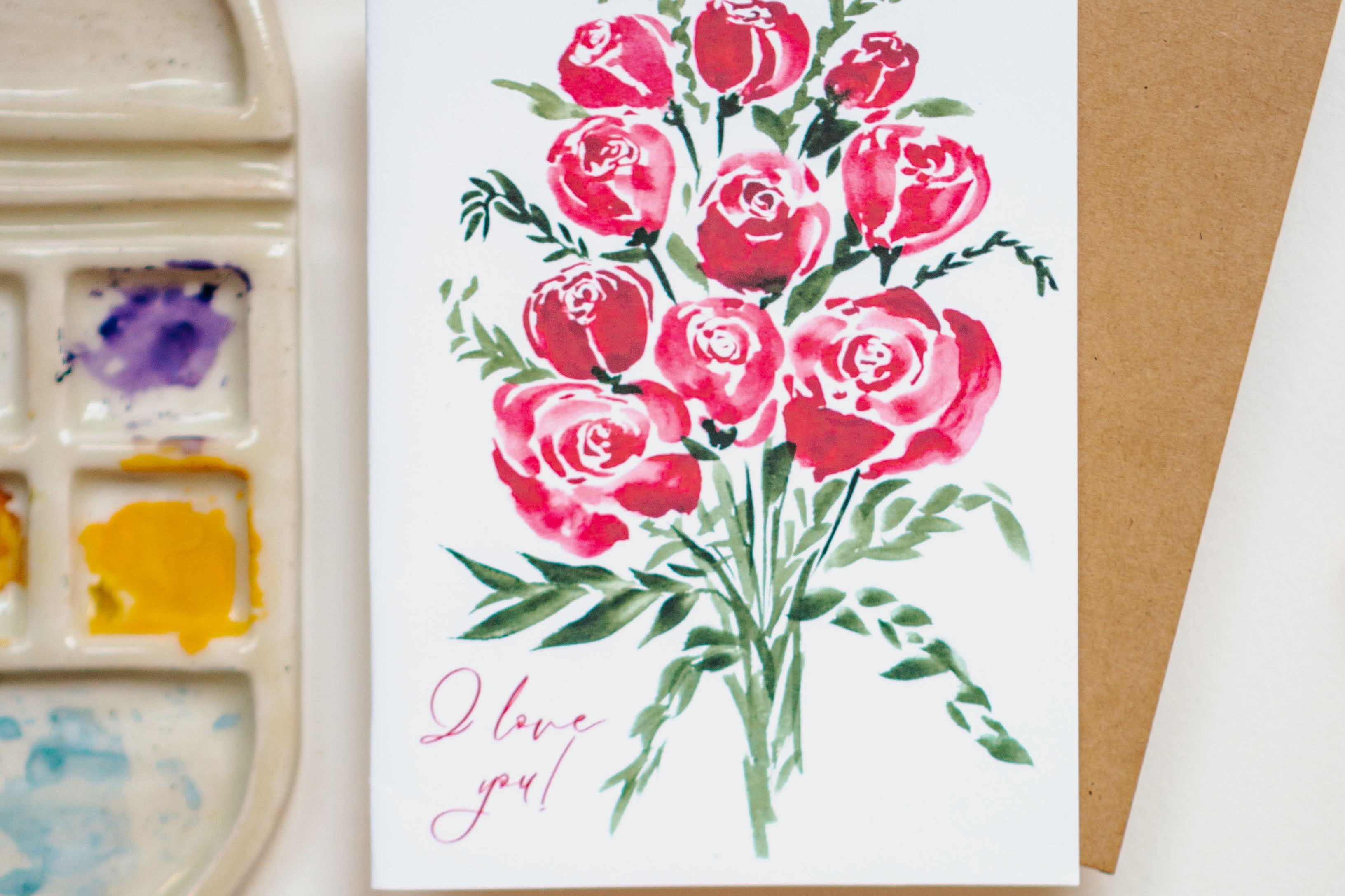 A white greeting card with red watercolor roses and "I love you!" written in thin, cursive script in the bottom left corner.