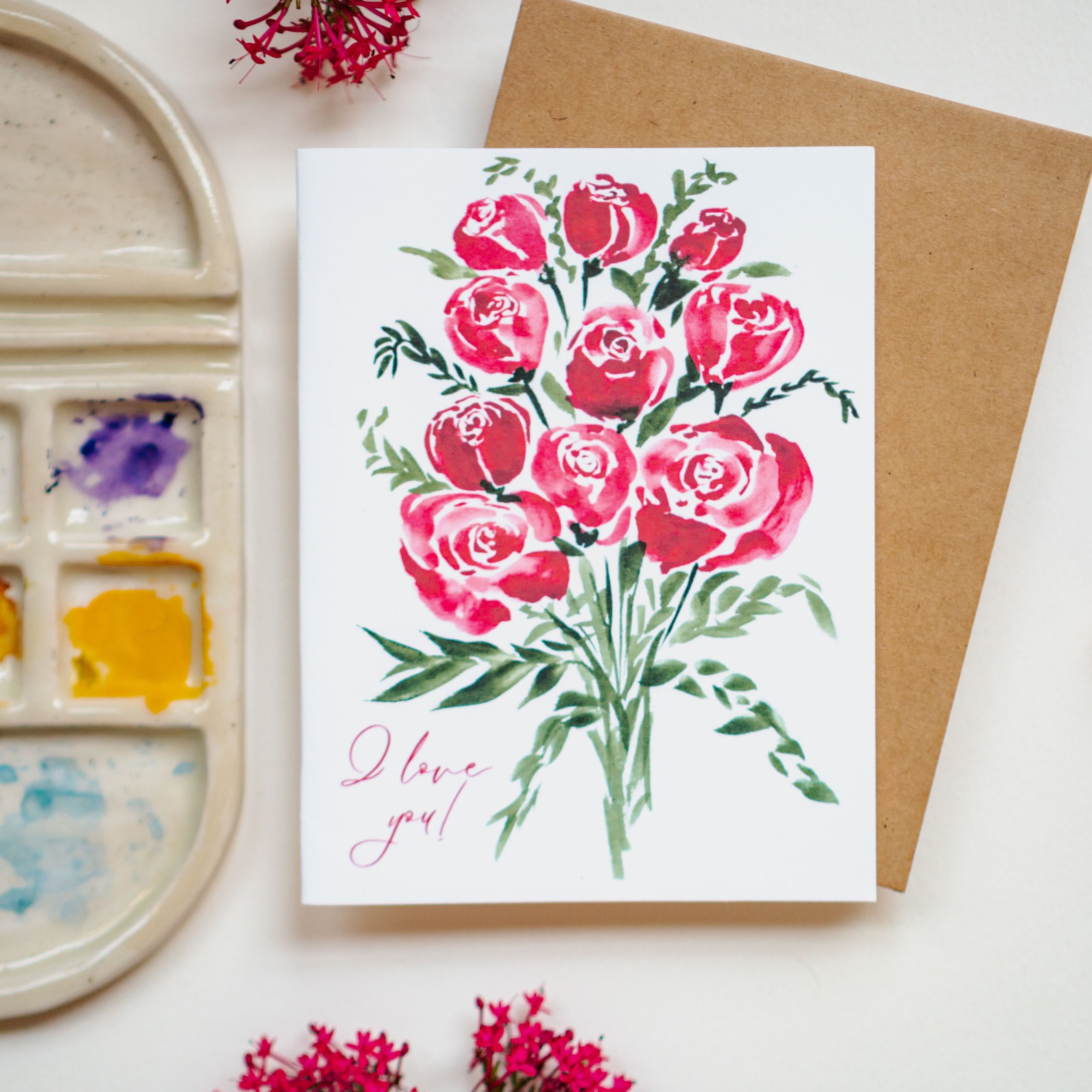 A white greeting card with red watercolor roses and "I love you!" written in thin, cursive script in the bottom left corner.
