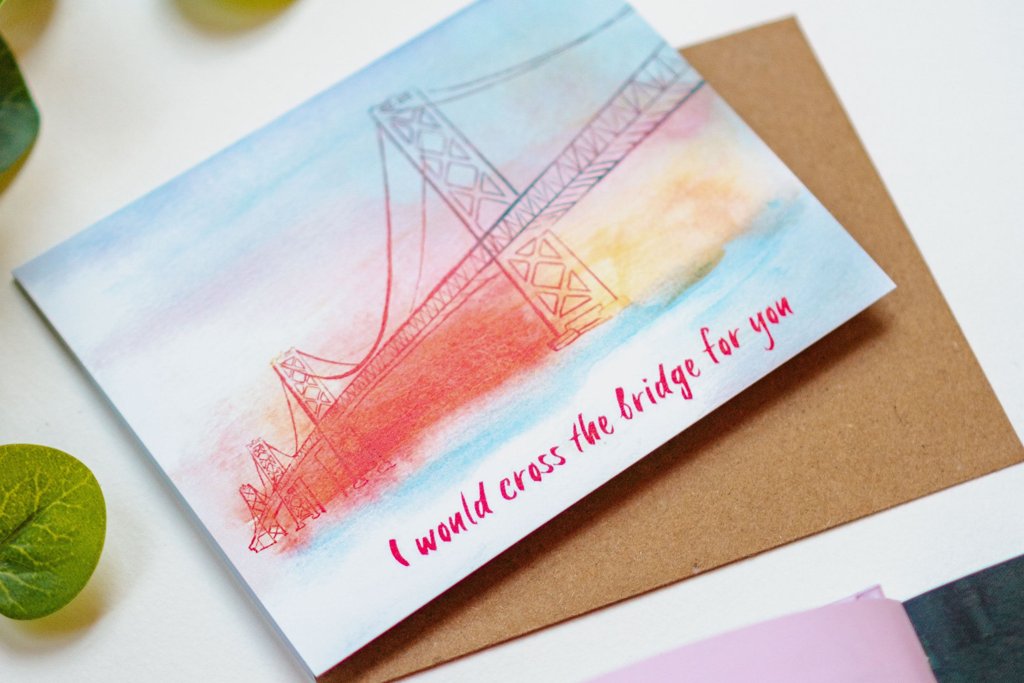 A card with a watercolor rendition of the San Francisco Bay Bridge on it, painted in oranges, yellows, and blues. The phrase "I would cross the bridge for you" is written on the bottom of the card.