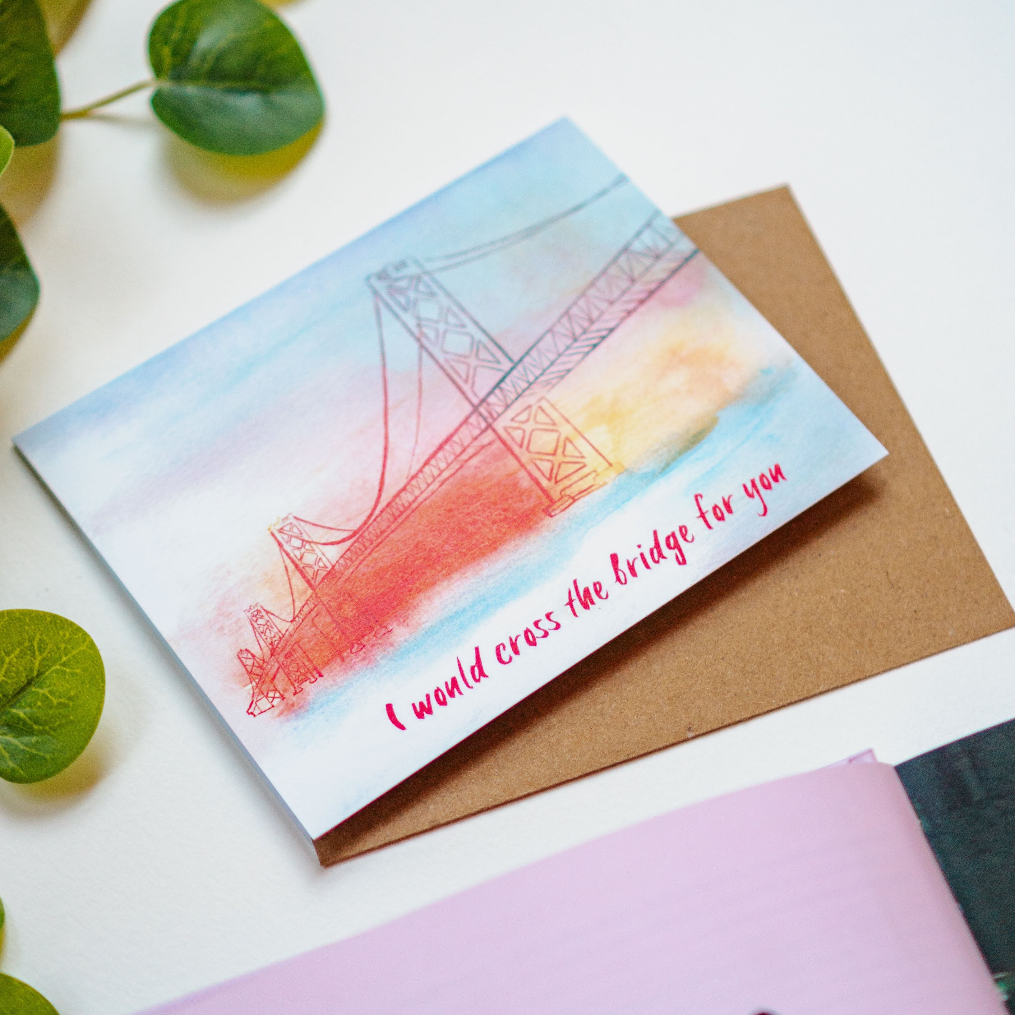 A card with a watercolor rendition of the San Francisco Bay Bridge on it, painted in oranges, yellows, and blues. The phrase "I would cross the bridge for you" is written on the bottom of the card.