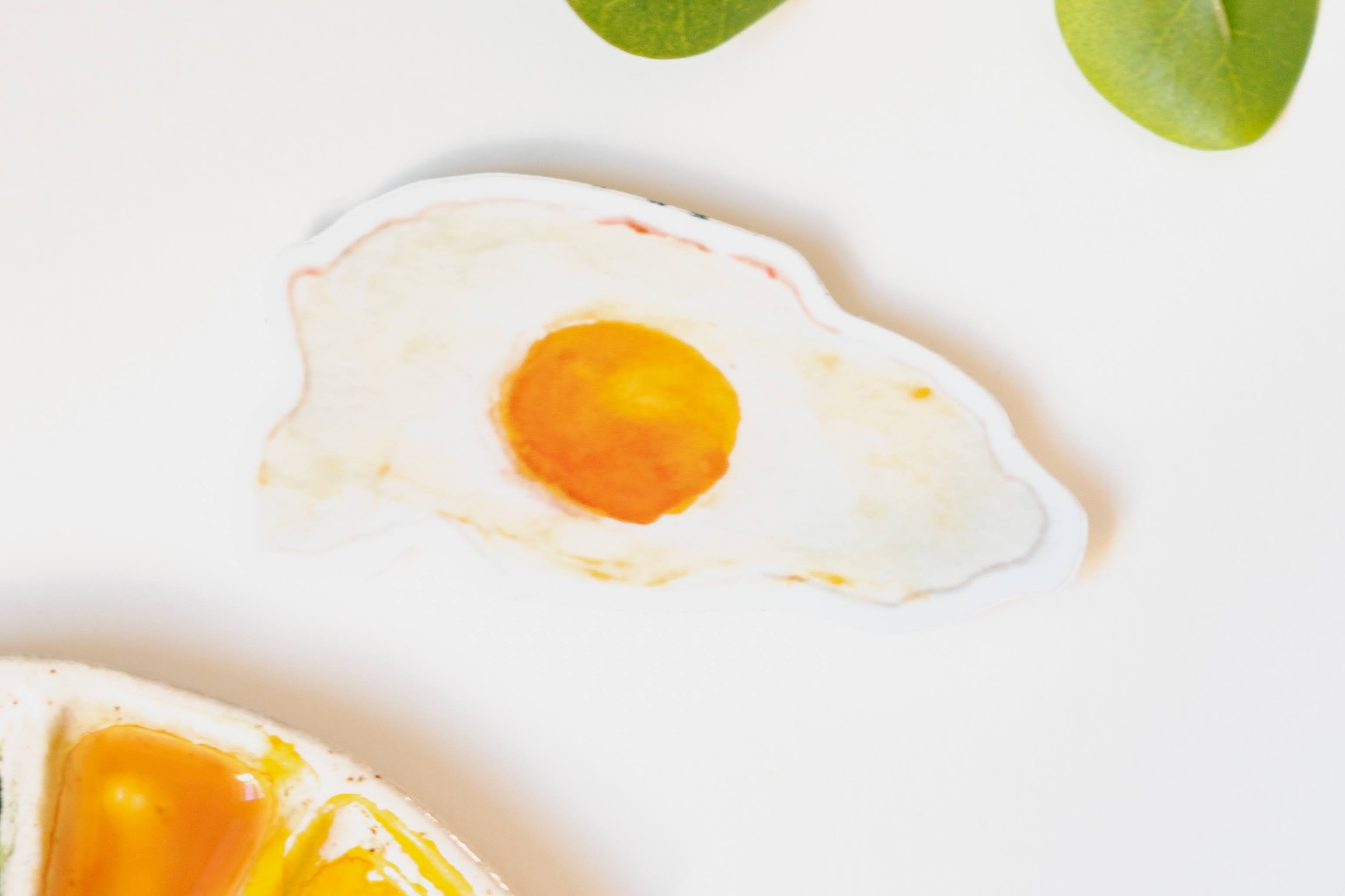 A narrow, white sticker of a fried, sunny side up egg with a rich yellow yolk and crispy edges.