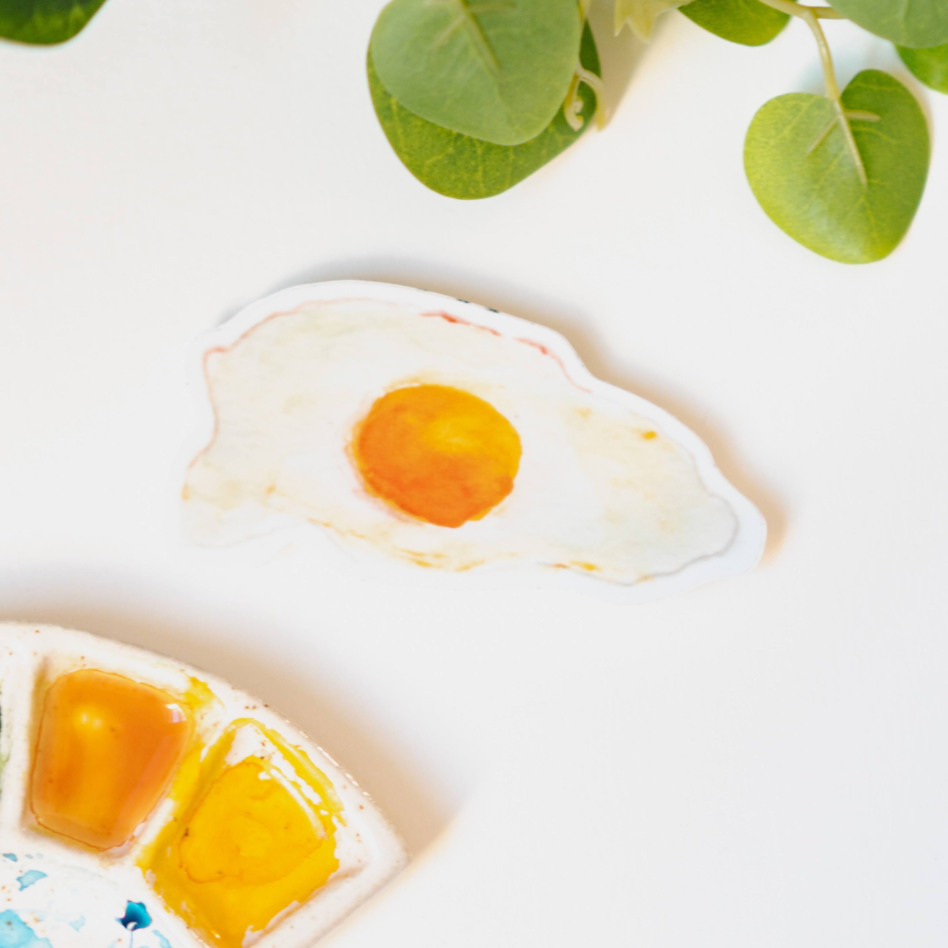 A narrow, white sticker of a fried, sunny side up egg with a rich yellow yolk and crispy edges.