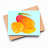 A light blue card with a red and orange mango on it. A cubed mango and a mango slice rests in front of a whole mango on the card.