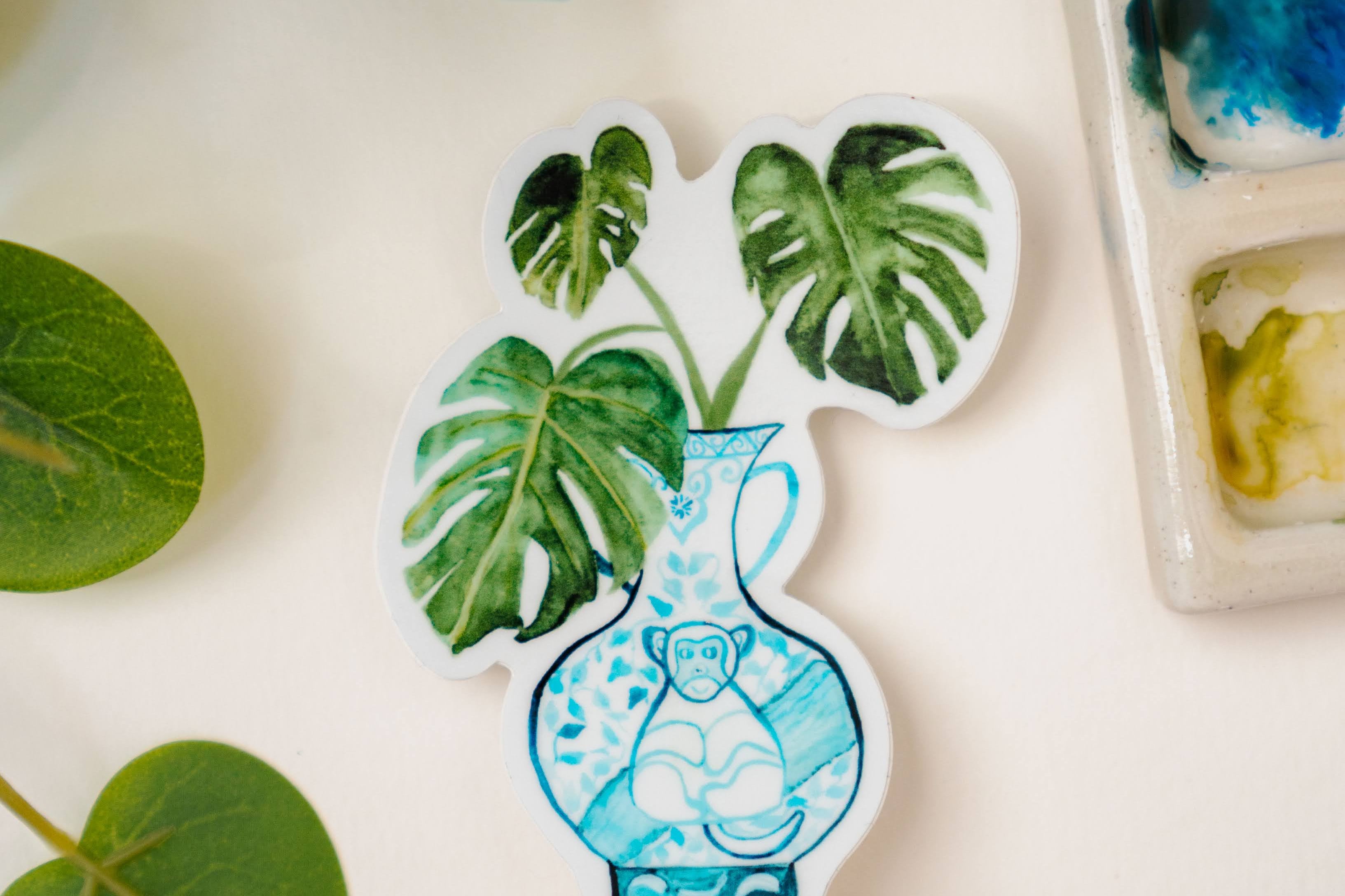 A sticker of a monstera plant in a blue and white porcelain vase, decorated with a monkey.