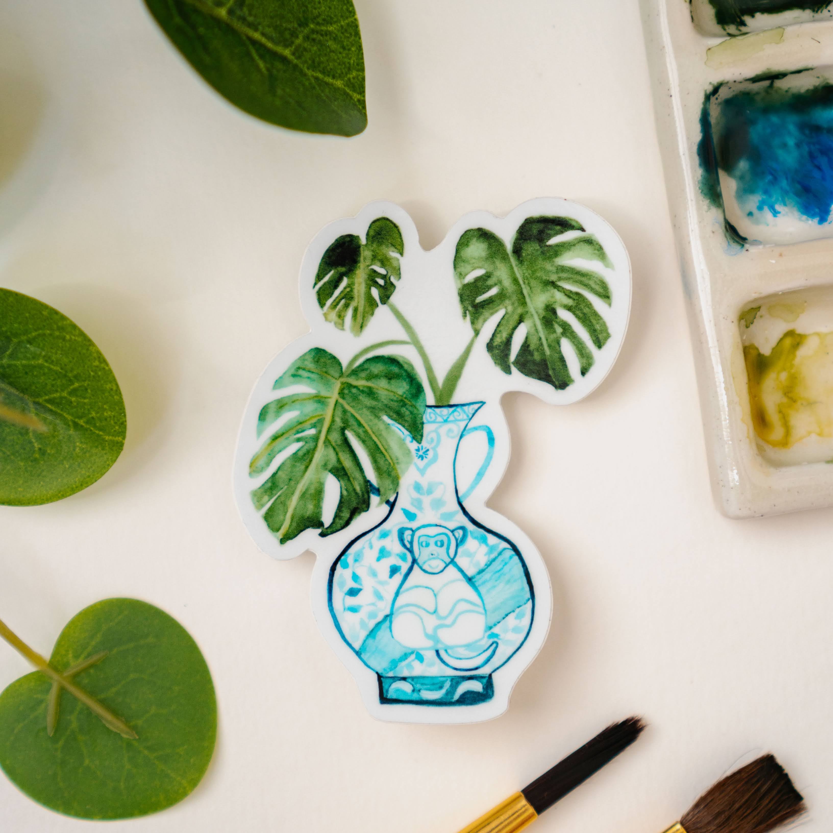 A sticker of a monstera plant in a blue and white porcelain vase, decorated with a monkey.