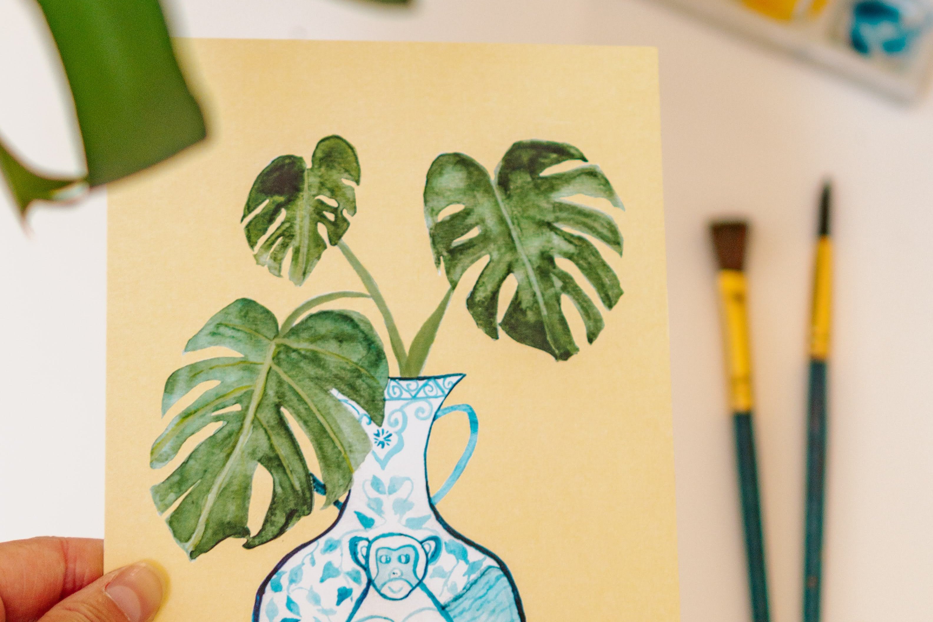 A print of a monstera plant in a blue and white porcelain vase, decorated with a monkey.