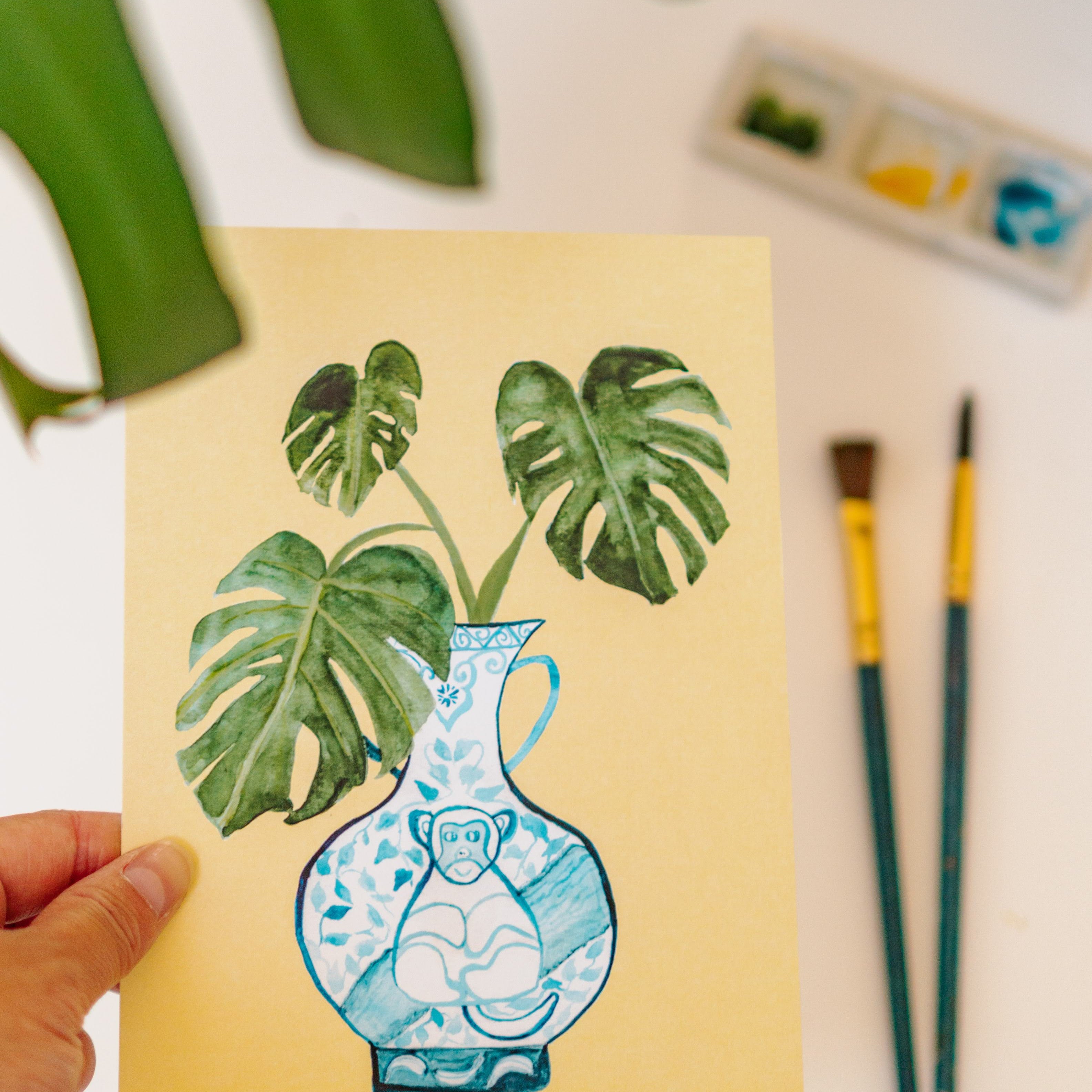 A print of a monstera plant in a blue and white porcelain vase, decorated with a monkey.