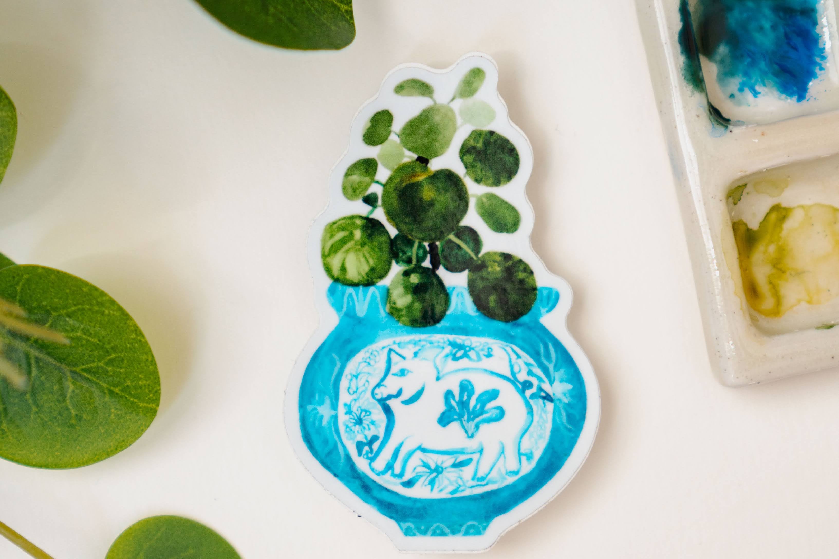 A sticker of a pilea plant in a blue and white porcelain vase, decorated with a pig.