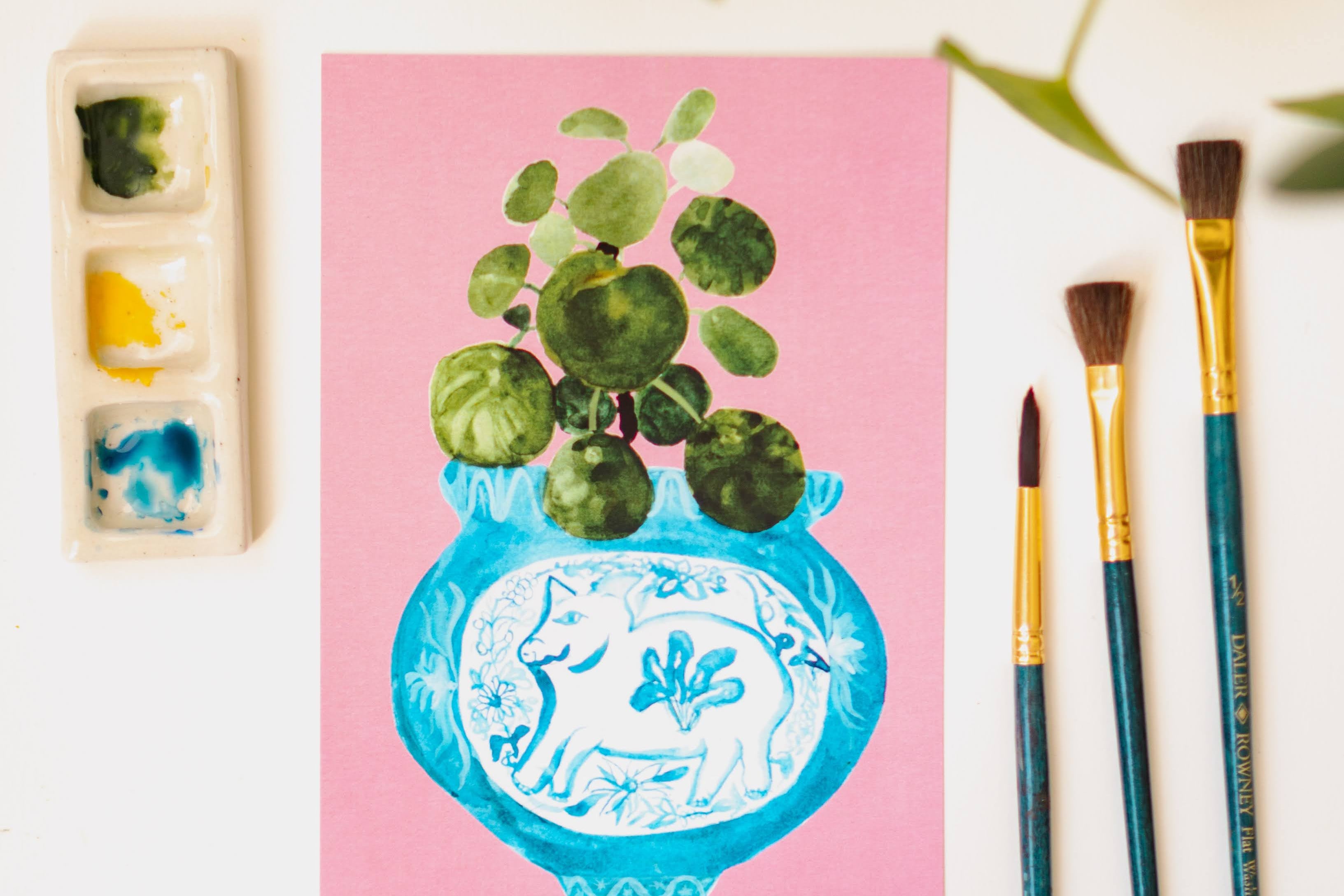 A print of a pilea plant in a blue and white porcelain vase, decorated with a pig.